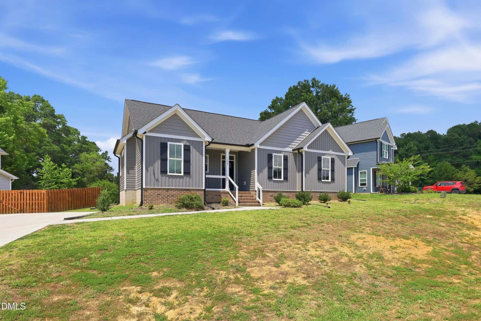 A single-story gray house with white trim, front porch, and brick foundation sits on a sloped lawn under a blue sky. A driveway and fenced backyard are visible, with trees and another house nearby.