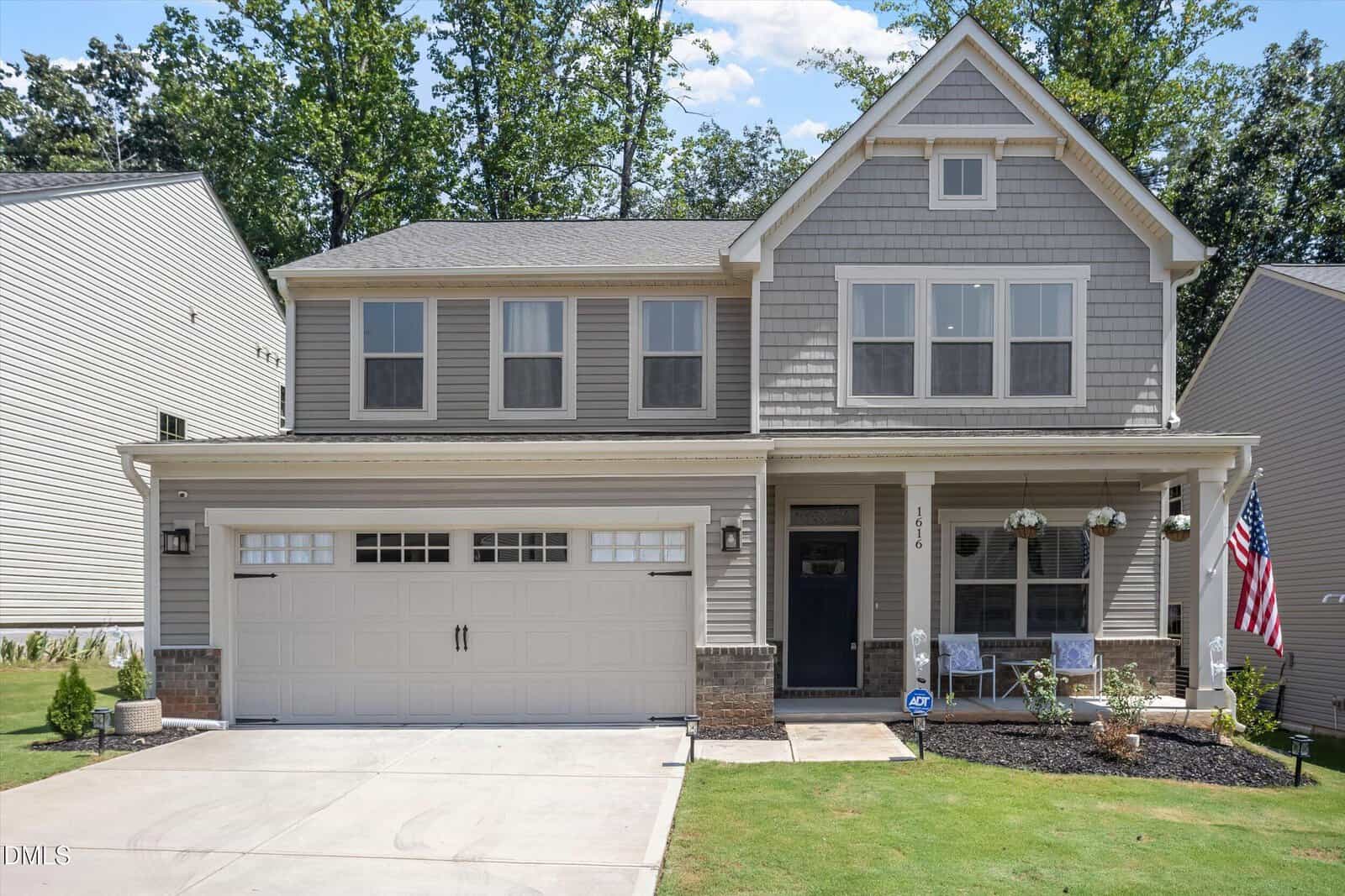 Two-story gray house with a double garage, front porch with chairs, American flag, hanging flower baskets, and a well-kept lawn. Located at 1616 Crimson Leaf Lane in Wake Forest, this home features trees and neighboring houses visible in the background.