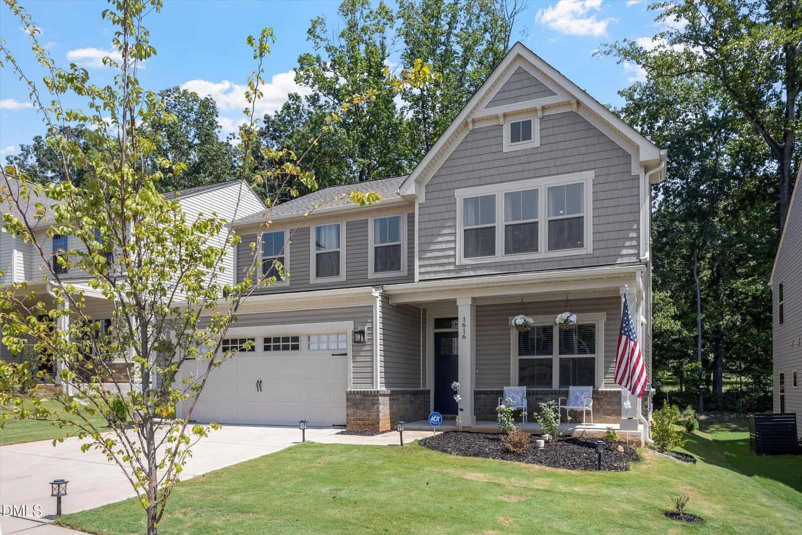Two-story suburban house with gray siding, two-car garage, and front porch. American flag and two chairs on porch. Well-kept lawn, young tree, and wooded background under a blue sky.