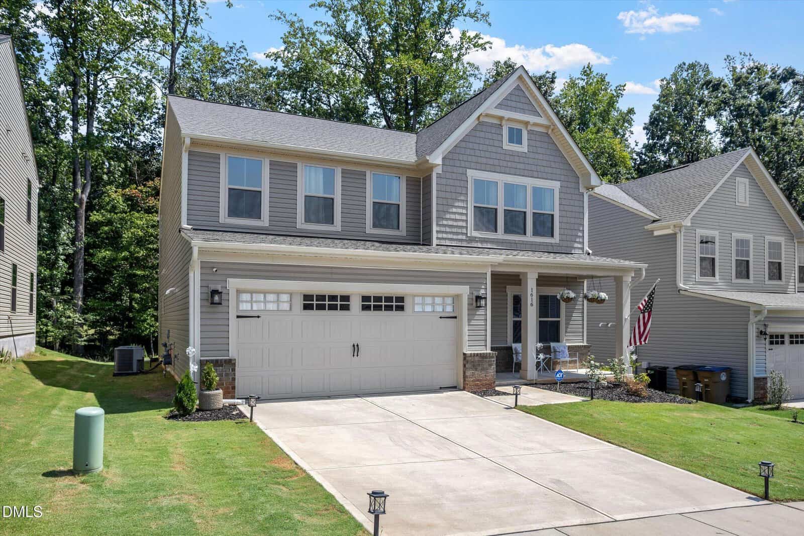 Two-story gray suburban house with attached two-car garage, neatly manicured lawn, American flag, and trees in the background on a sunny day.