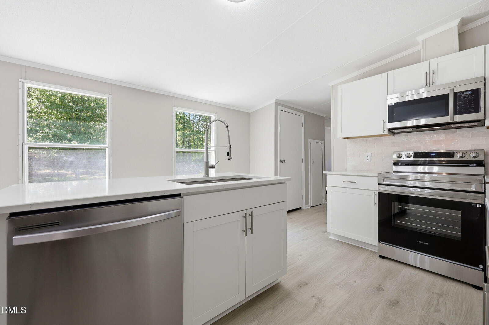 Modern kitchen with white cabinets, stainless steel appliances, a built-in microwave, dishwasher, electric stove, central island with sink, and large windows letting in natural light. Light wood flooring throughout.