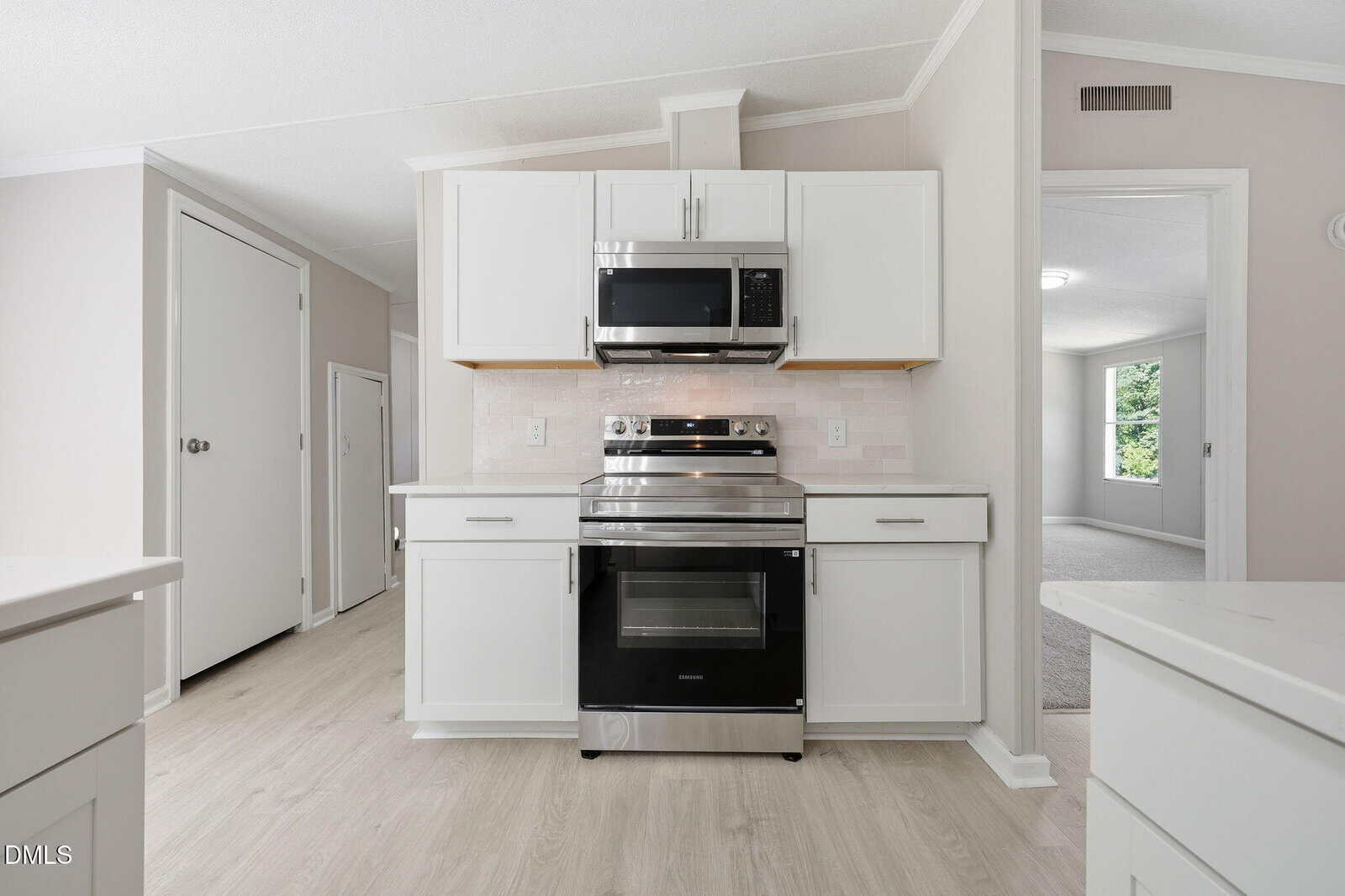 Modern kitchen with white cabinets, stainless steel oven and microwave, light wood flooring, and a neutral backsplash. An open doorway leads to a carpeted room with a window and greenery visible outside.