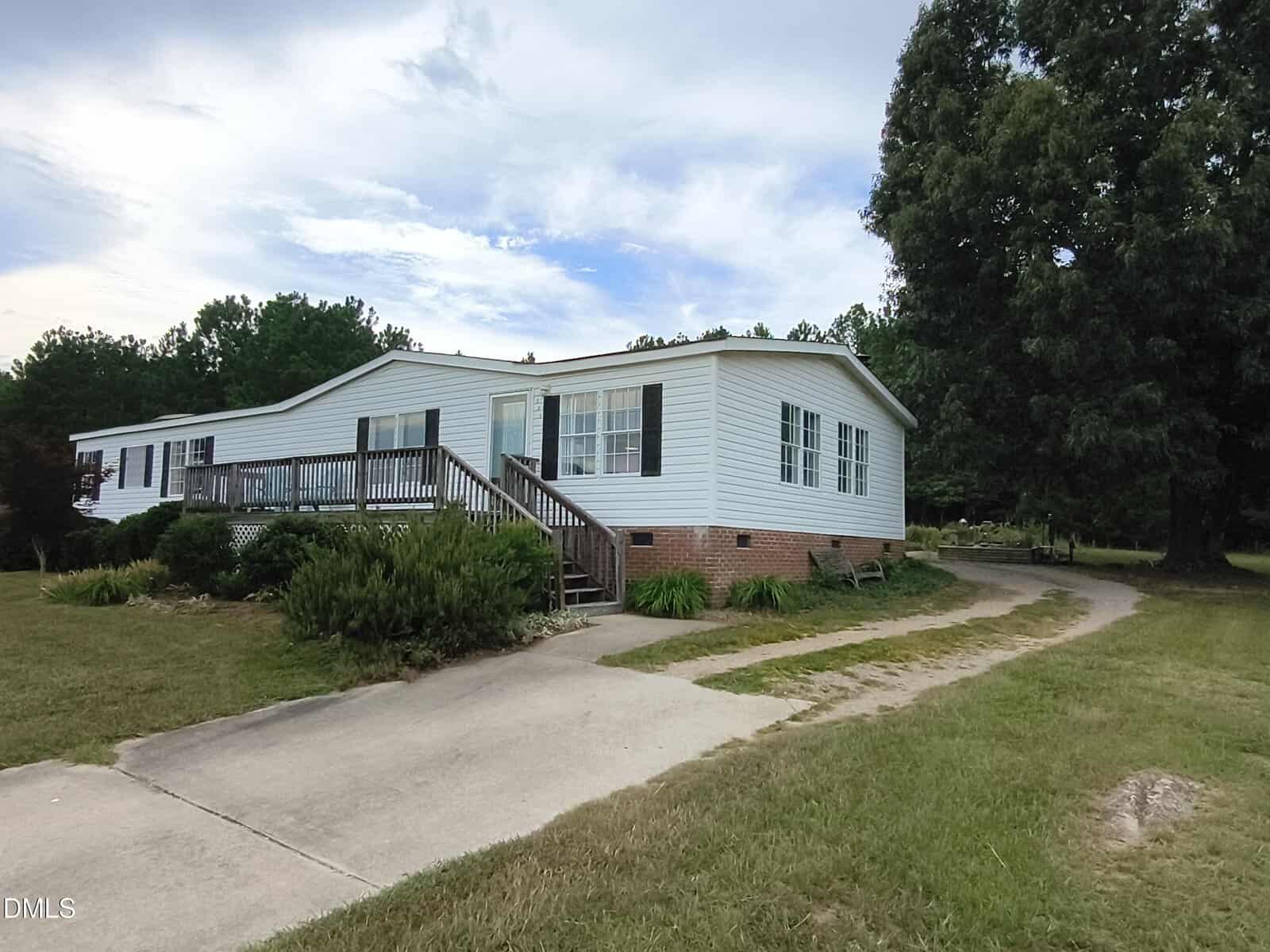 White single-story house with large windows, a front porch with steps, and a curved driveway surrounded by grass, bushes, and trees under a partly cloudy sky.