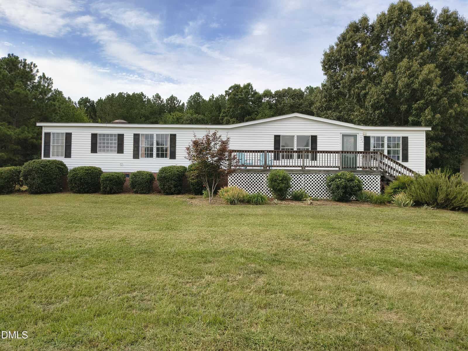 Single-story white house with black shutters, front porch with railings, stairs, manicured bushes, small trees, and large green lawn at 125 N North Ridge Drive in Louisburg. Background features tall trees under a partly cloudy sky.