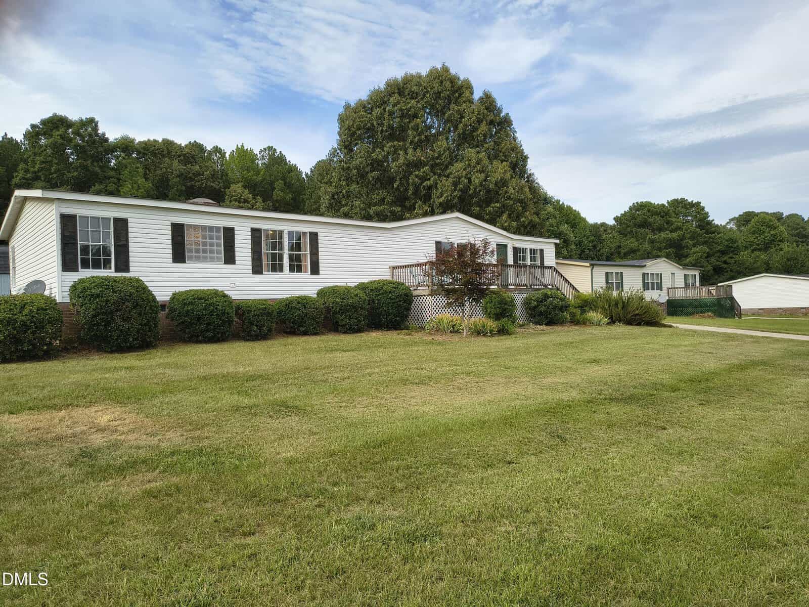 A white, single-story manufactured home with black shutters sits on a large grassy lawn, surrounded by neatly trimmed bushes and trees under a partly cloudy sky.