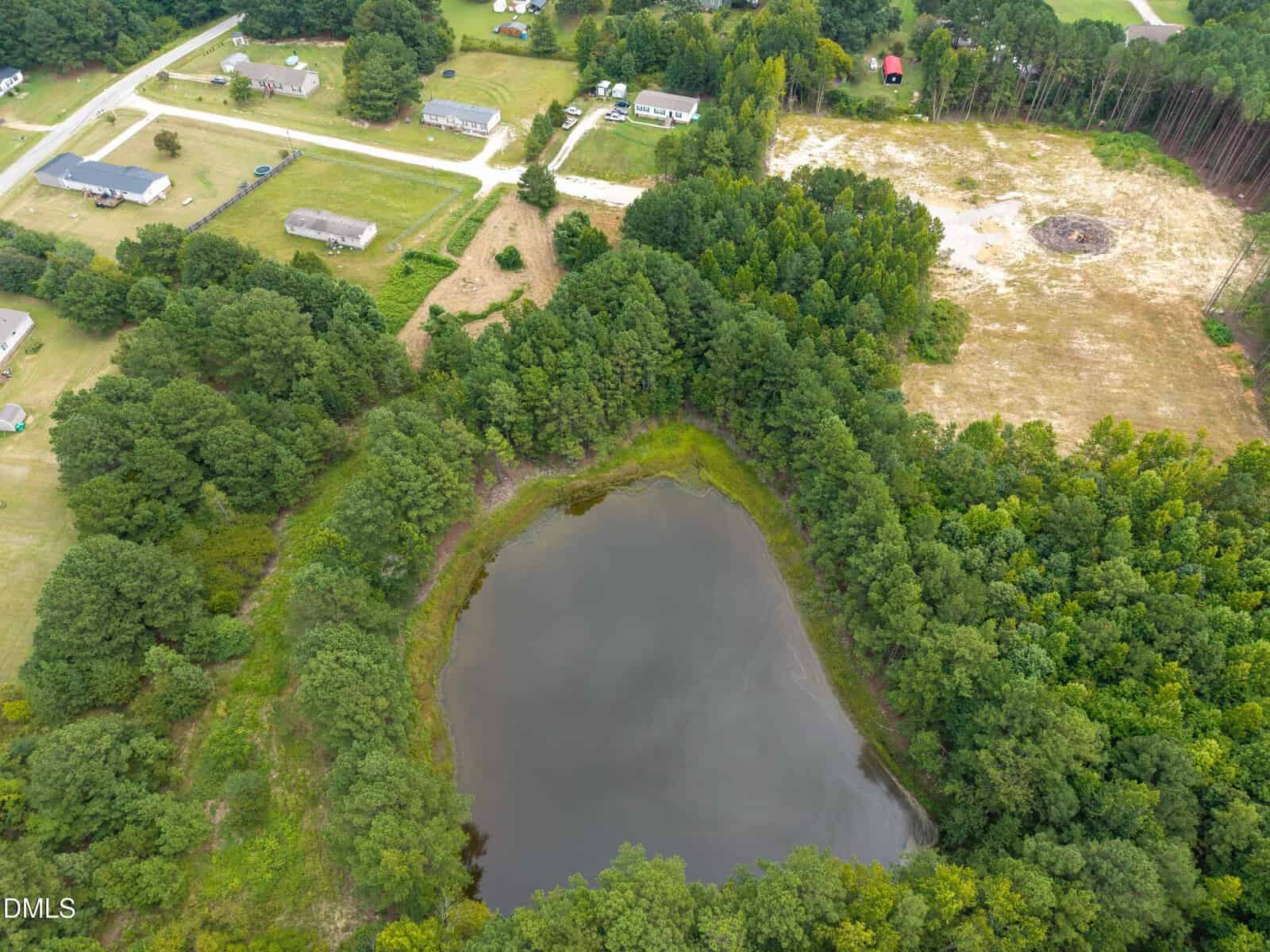 Aerial view of a small pond surrounded by dense trees, with nearby houses, roads, and open grassy areas in a rural Henderson neighborhood near 14r Grove Park Lane.