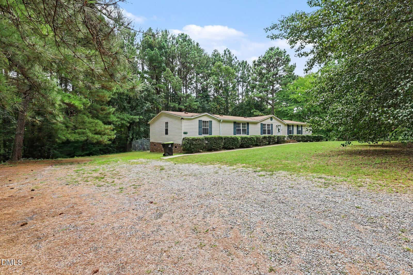 A beige manufactured home with green shutters sits on a grassy lawn surrounded by tall trees, with a gravel driveway in the foreground under a blue sky.