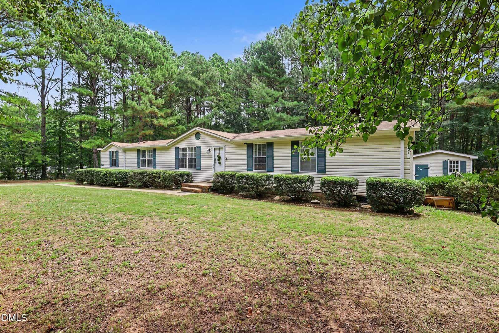 Nestled at 3542 Charlies Court in Wake Forest, this single-story white house features charming blue shutters, small front steps, manicured bushes, and a grassy lawn, all surrounded by tall pine trees under a partly cloudy sky.