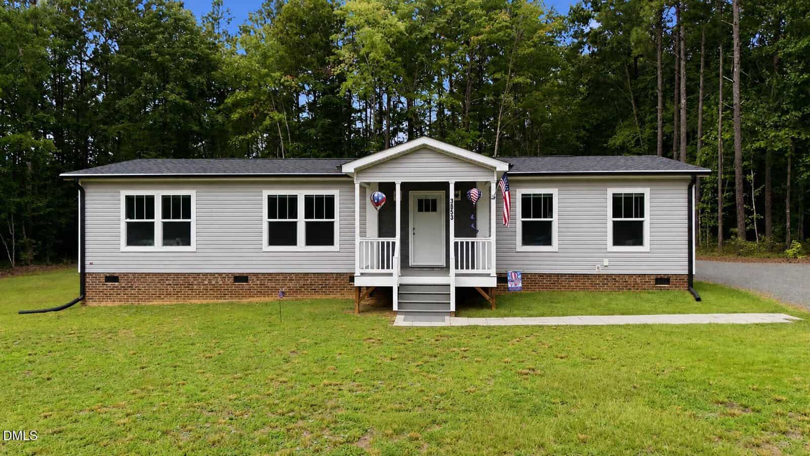 Single-story house with light gray siding, brick foundation, and small front porch located at 3053 Sterl Carrington Road, Oxford. American flags and patriotic decorations hang near the entrance. The home is surrounded by grass and trees.