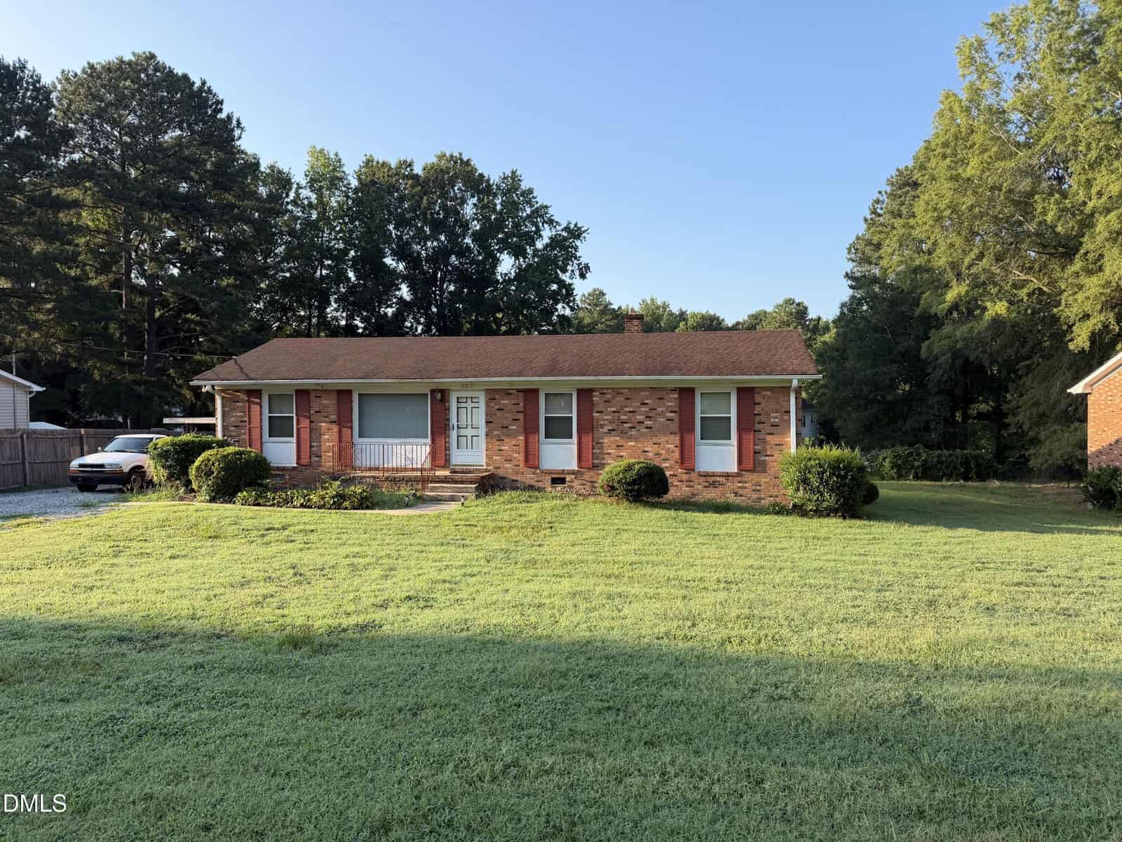 Single-story brick house with a brown roof, red shutters, and a large covered front porch, set on a grassy lawn with bushes, trees, and a clear blue sky in the background—perfectly situated at 317 Aiken Avenue in the charming community of Creedmoor.