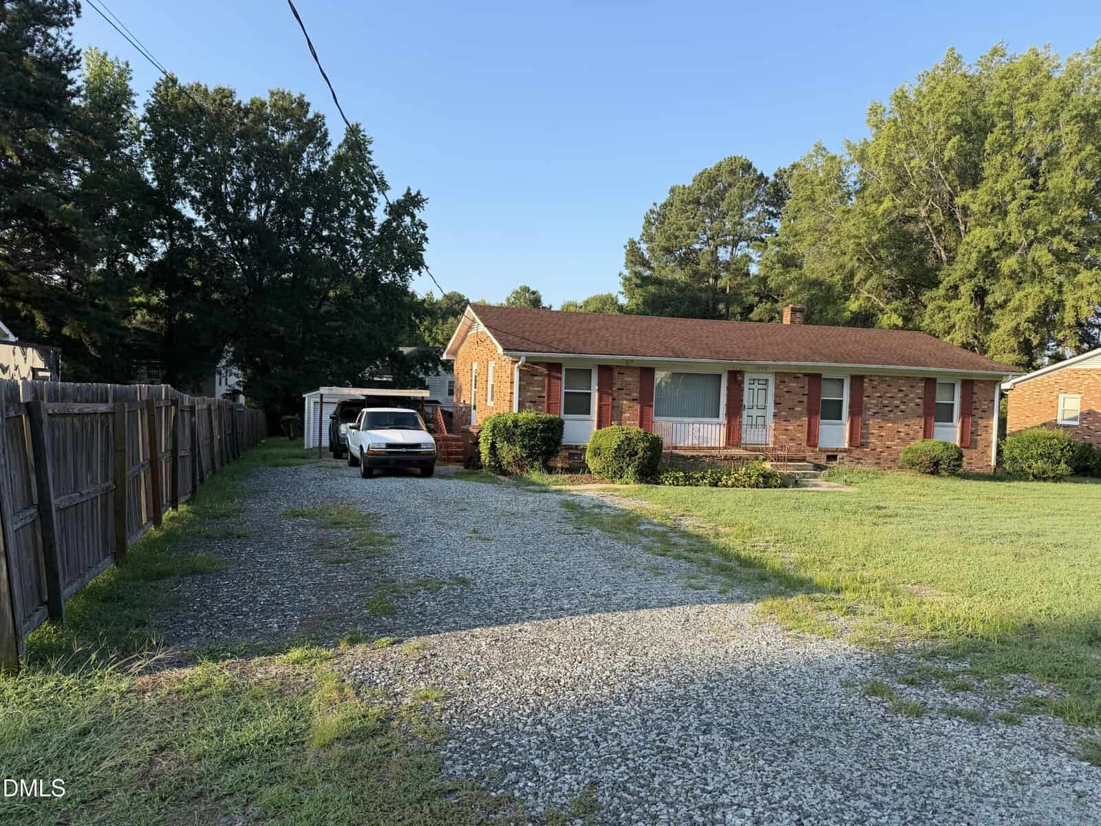 A one-story brick house with a red roof, a gravel driveway, a parked white car, green grass, bushes, and trees under a clear blue sky.