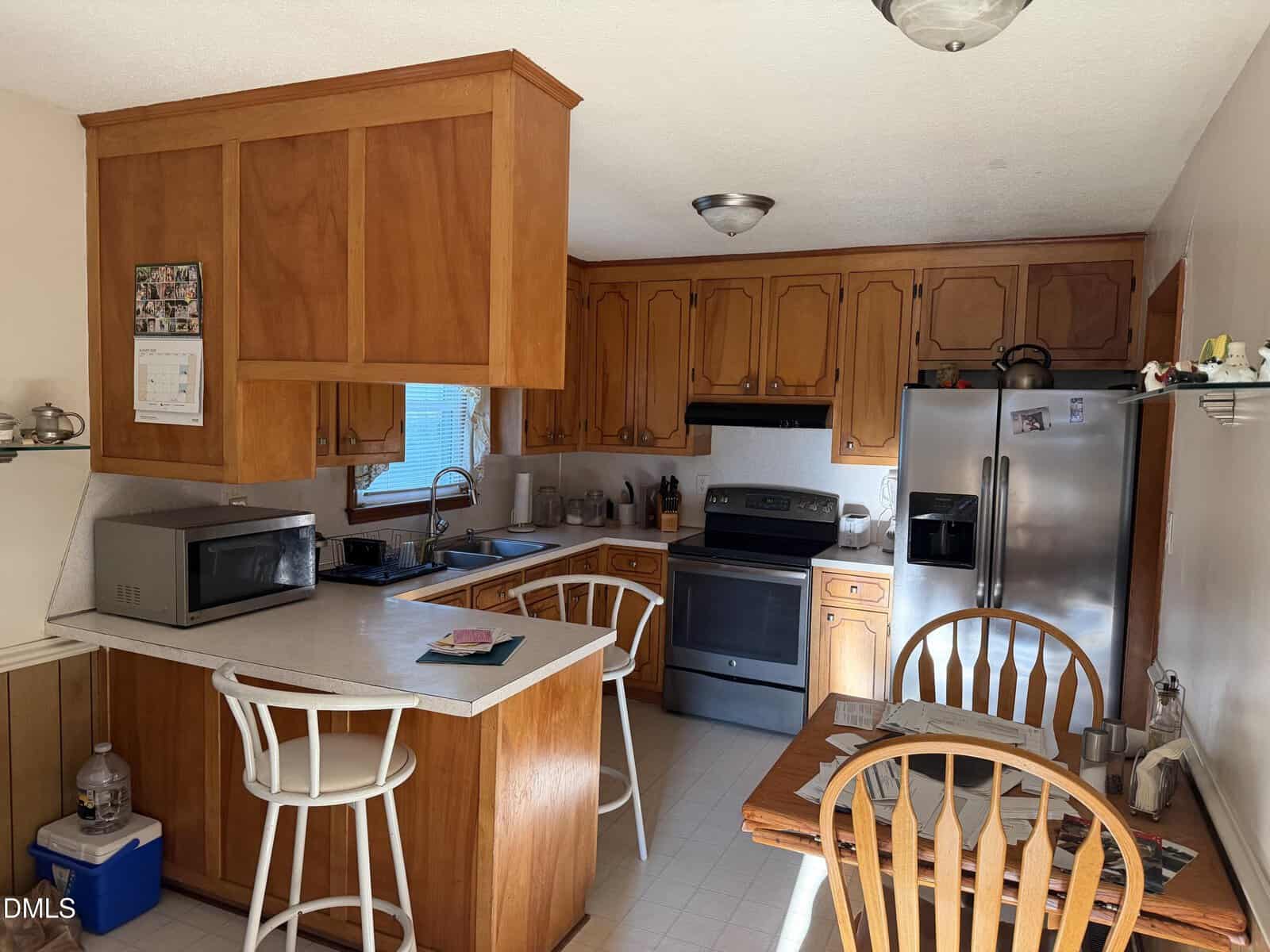 A sunlit kitchen with wood cabinets, stainless steel appliances, an island with stools, and a small dining table set for two. Various kitchen items are on counters and shelves.