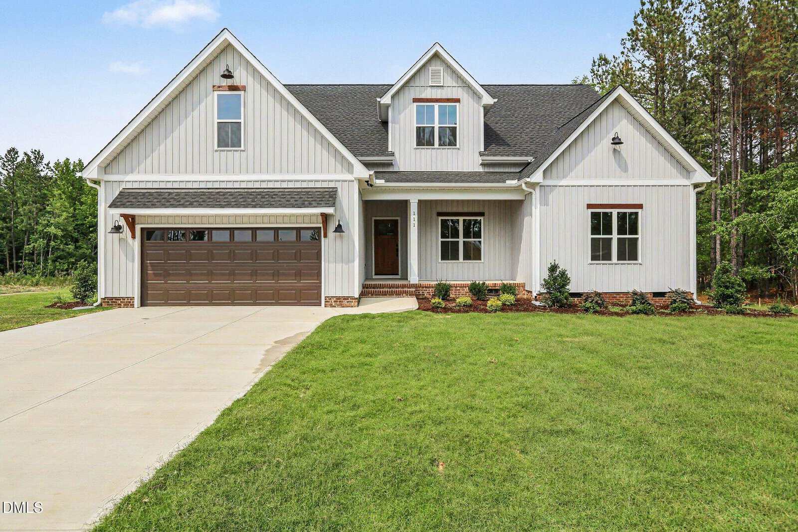 Modern white house with dark roof, two dormer windows, a double garage, and a large driveway. Green lawn and landscaped shrubs in front add to the curb appeal of 111 Stonepond Drive, Oxford, with trees in the background under a blue sky.