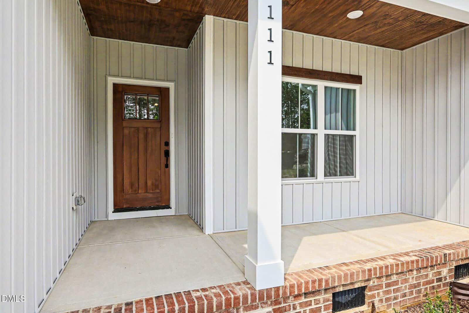 A covered front porch with a wooden door, white vertical siding, white column labeled “111,” a double window, and a brick base. The porch has a concrete floor and wooden ceiling accents.