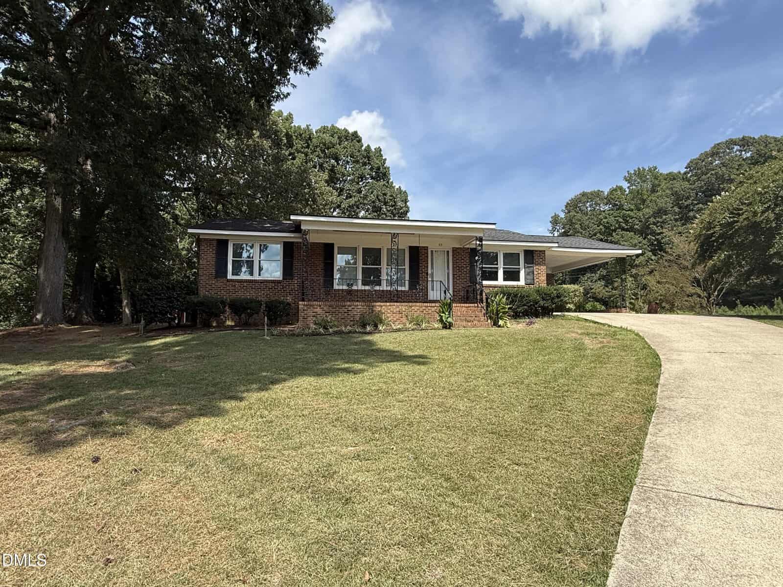 Single-story brick house with dark shutters, large front windows, and a sloped driveway, set on a grassy lawn bordered by trees under a blue sky with scattered clouds—conveniently located at 65 Bunn Avenue in the charming town of Zebulon.