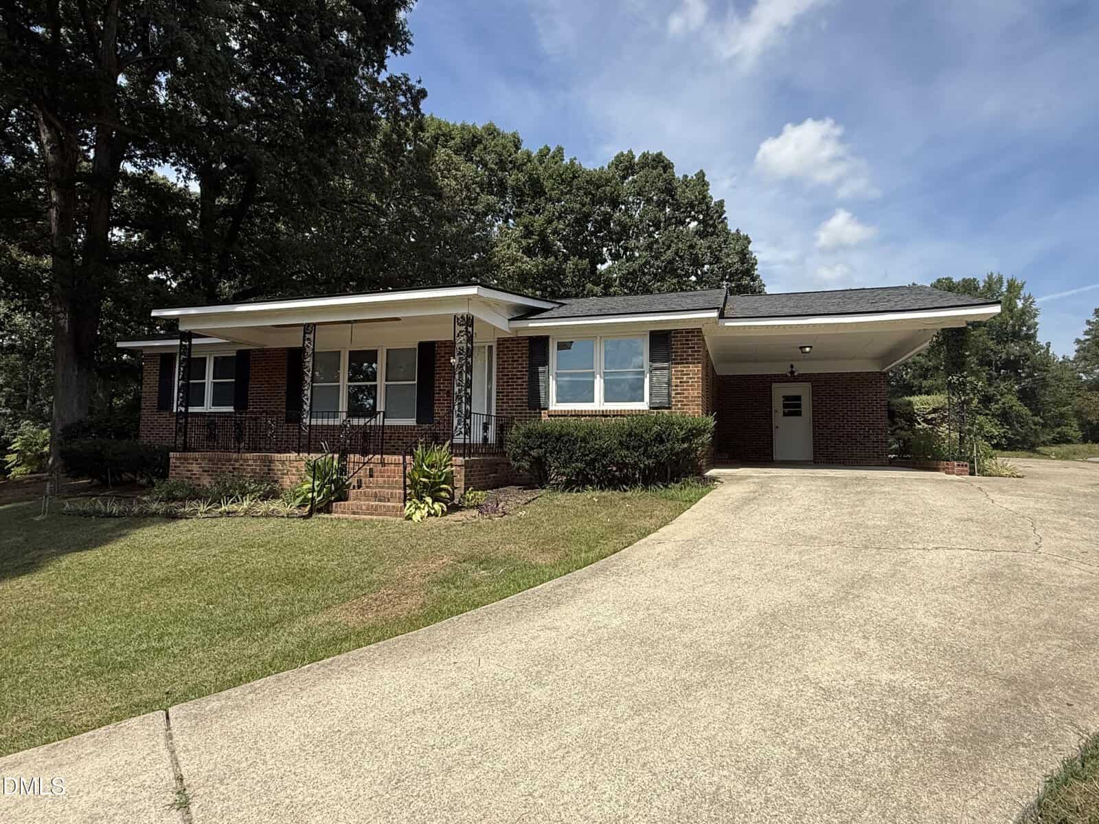 Single-story brick house with a covered carport, front porch with black railings, steps leading to the entrance, and well-kept lawn. Large trees and bushes surround the home under a blue sky with clouds.