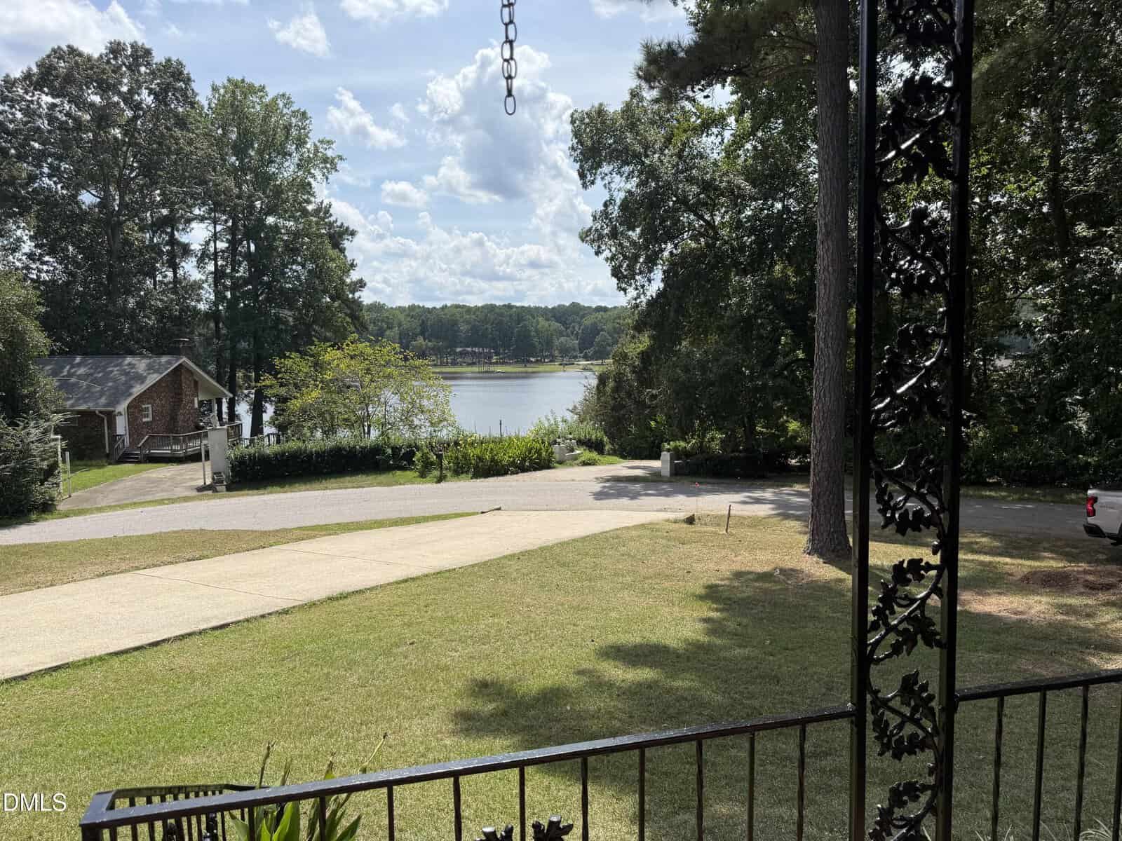 View from a porch overlooking a grassy yard, driveway, road, and a small lake surrounded by trees, with a partly cloudy sky and a house visible on the left near the water.