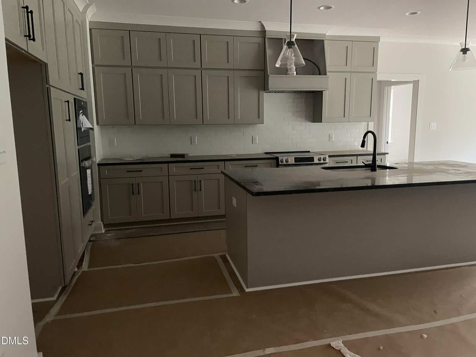 Modern kitchen with gray cabinets, black countertops, a large island with sink, stainless steel stove, and white subway tile backsplash. Floor is covered with protective brown paper. Room is well-lit with pendant lights.
