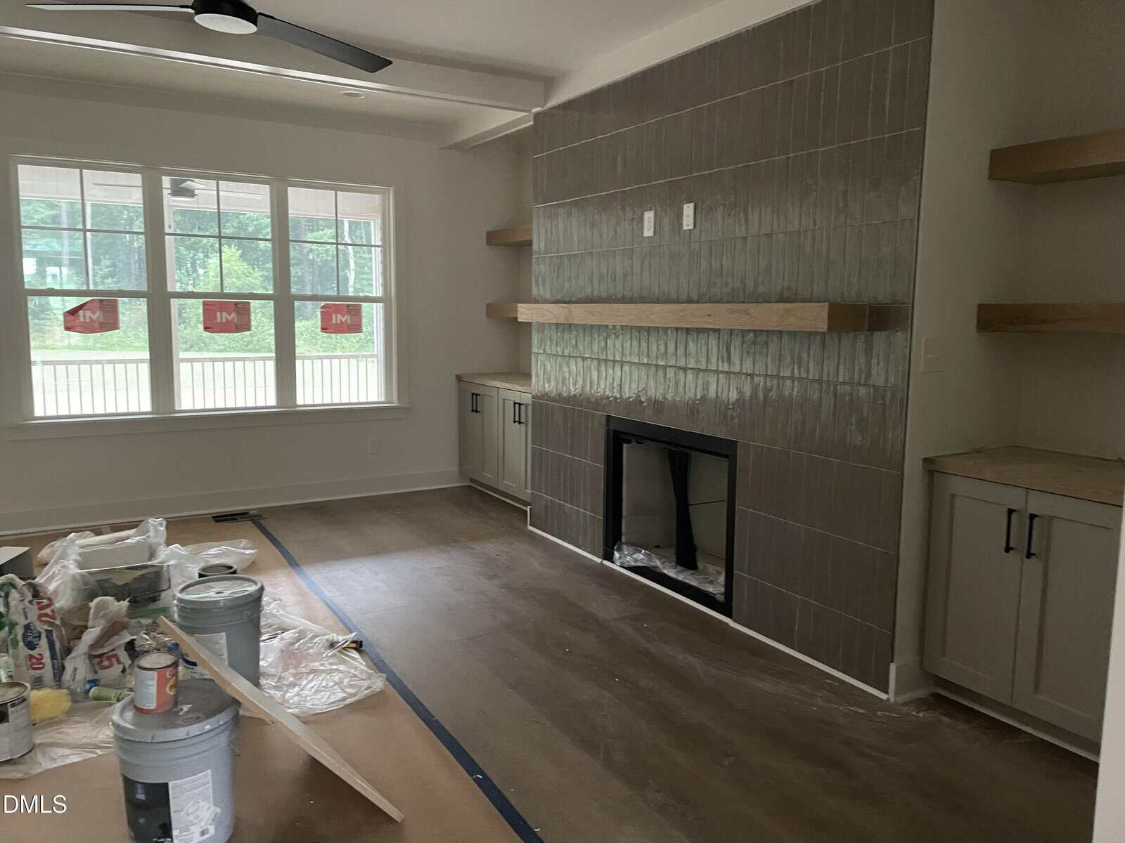 A living room under construction with wood floors, tile fireplace, floating shelves, cabinets, and large window. Paint cans and supplies are on a plastic-covered floor. Light streams in through the window.