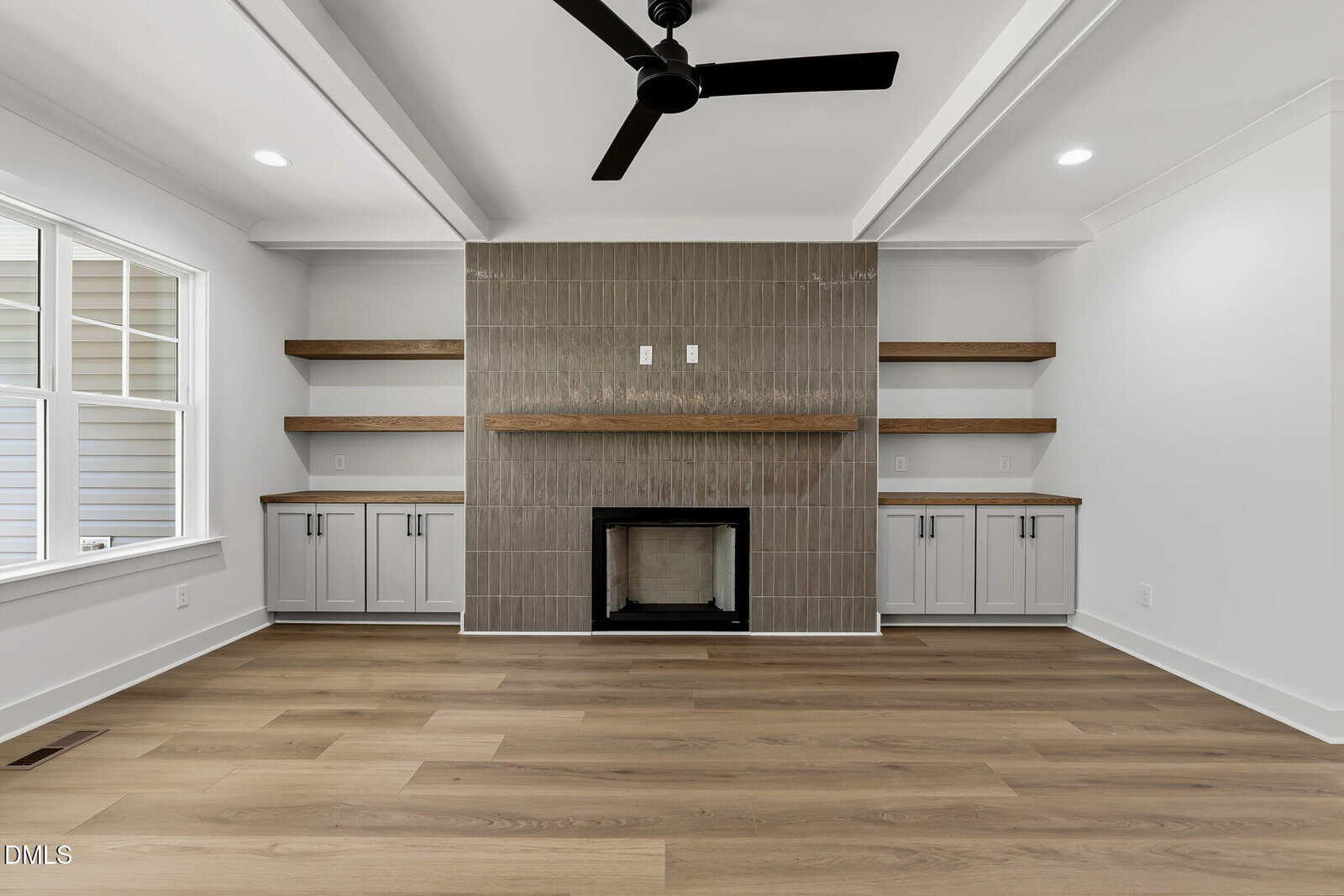 A modern living room with light wood floors, a tiled fireplace, floating wood shelves, white built-in cabinets, a ceiling fan, and a large window on the left wall.