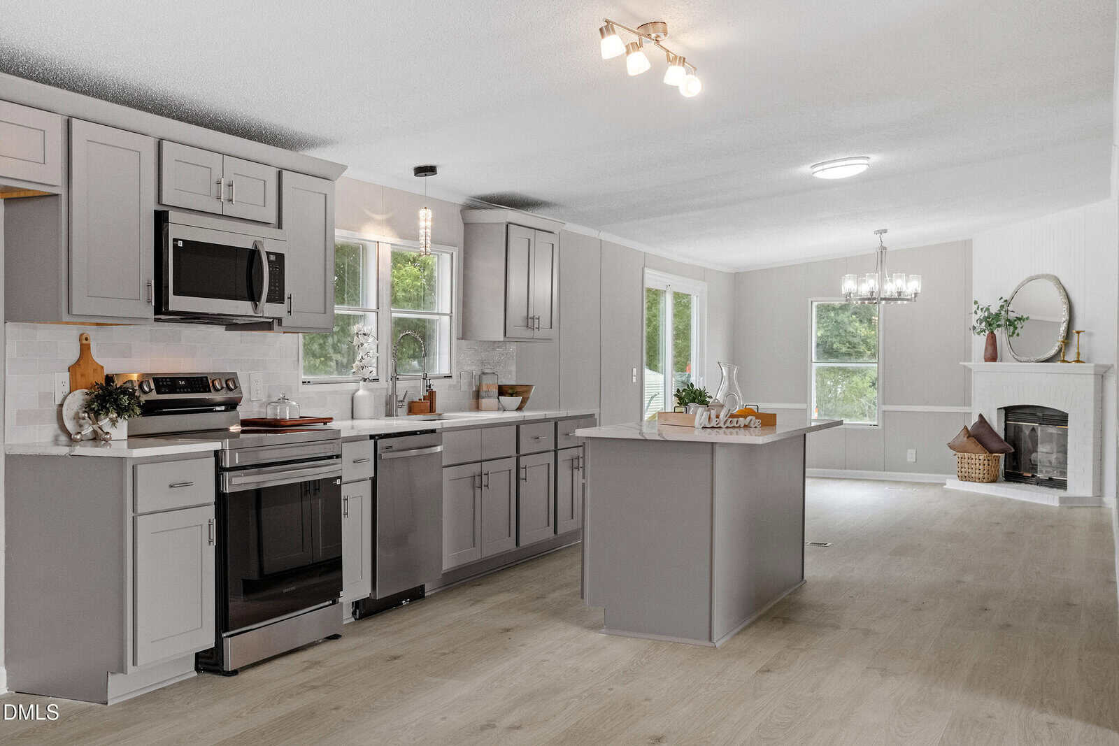 Modern kitchen with gray cabinets, stainless steel appliances, a center island, and light wood floors. In the background, a living area with a fireplace and large windows letting in natural light.