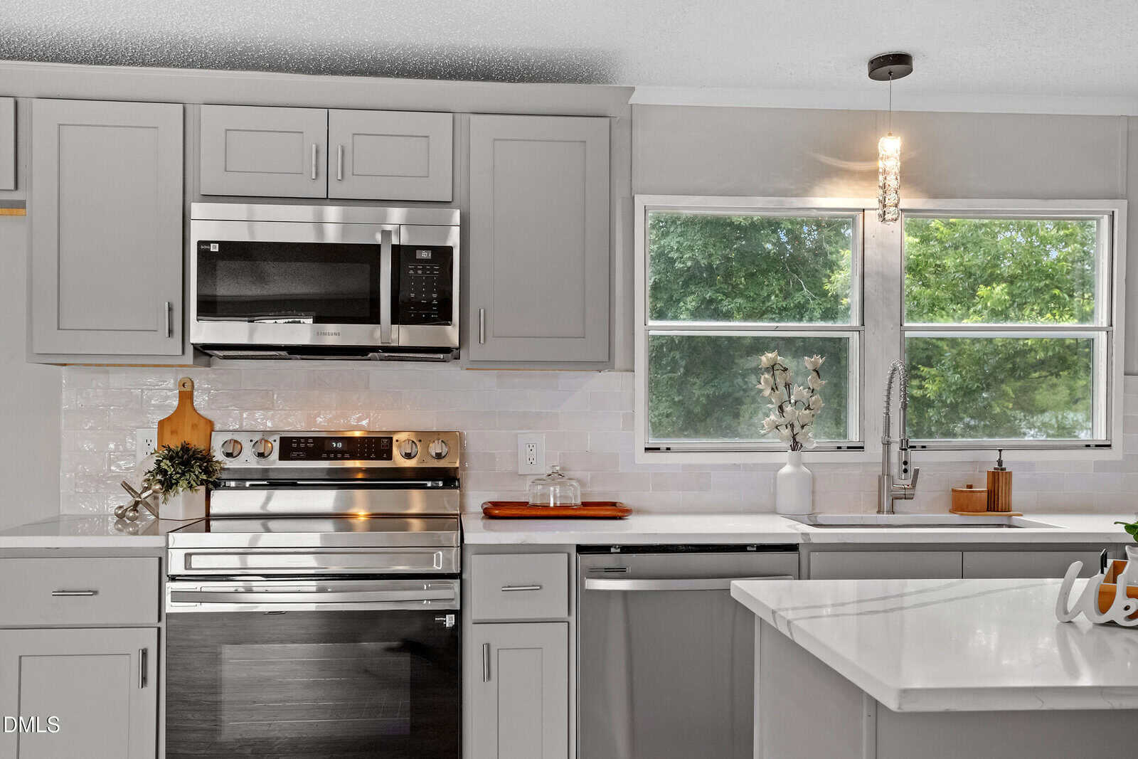Modern kitchen with gray cabinets, stainless steel appliances, double oven, and a microwave. A window is above the sink, letting in natural light. White countertops and backsplash give a clean, contemporary look.
