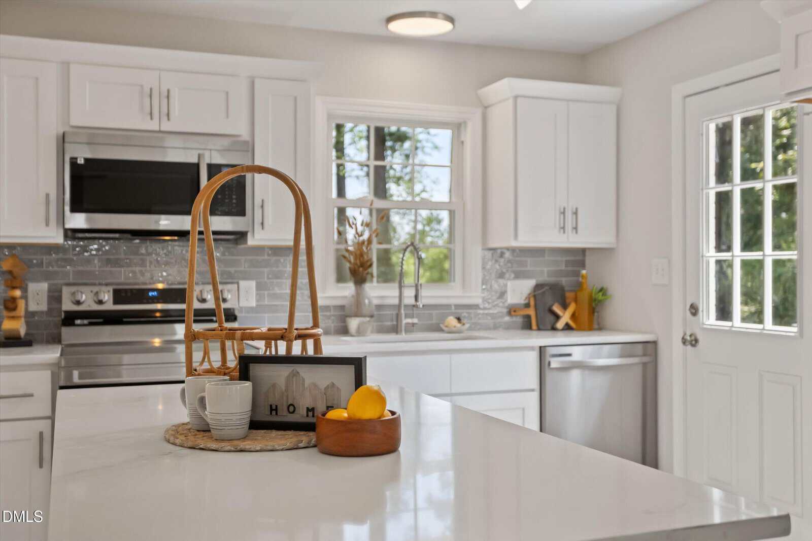 Bright, modern kitchen with white cabinets, stainless steel appliances, and a large island. A basket tray with mugs, a lemon, and a small “Home” sign sit on the island. Sunlight streams in through a window and glass door.