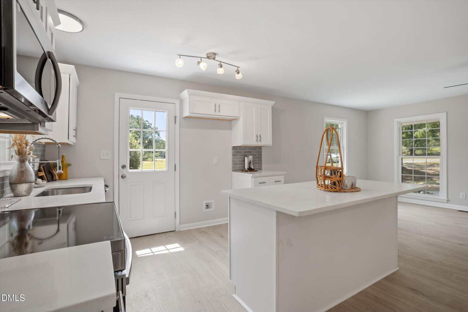 Bright modern kitchen with white cabinets, stainless steel appliances, an island with a wicker tray, light wood floors, and large windows letting in natural light. A door opens to a grassy outdoor area.