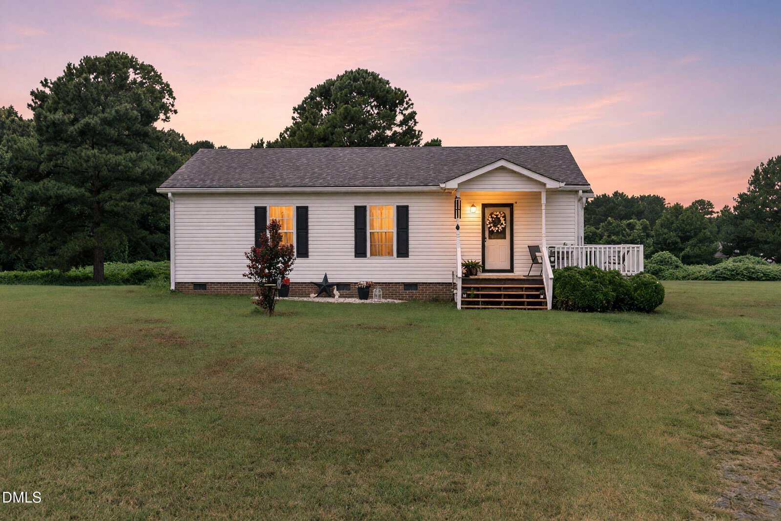 A small white house with black shutters, a front porch, and a wreath on the door sits on a large grassy lawn at sunset with trees in the background—an inviting scene found at 135 Stillmeadow Drive, Louisburg.