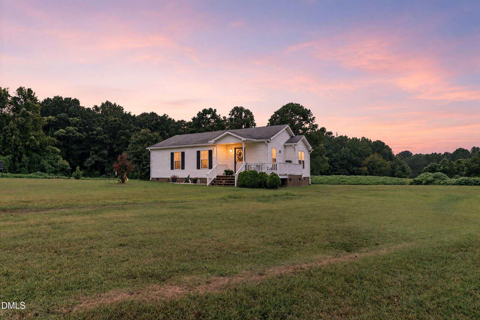 A small, white house with a porch stands on a large, open grassy lawn at sunset, surrounded by trees under a pink and blue sky. Warm lights glow from the windows, creating a cozy atmosphere.