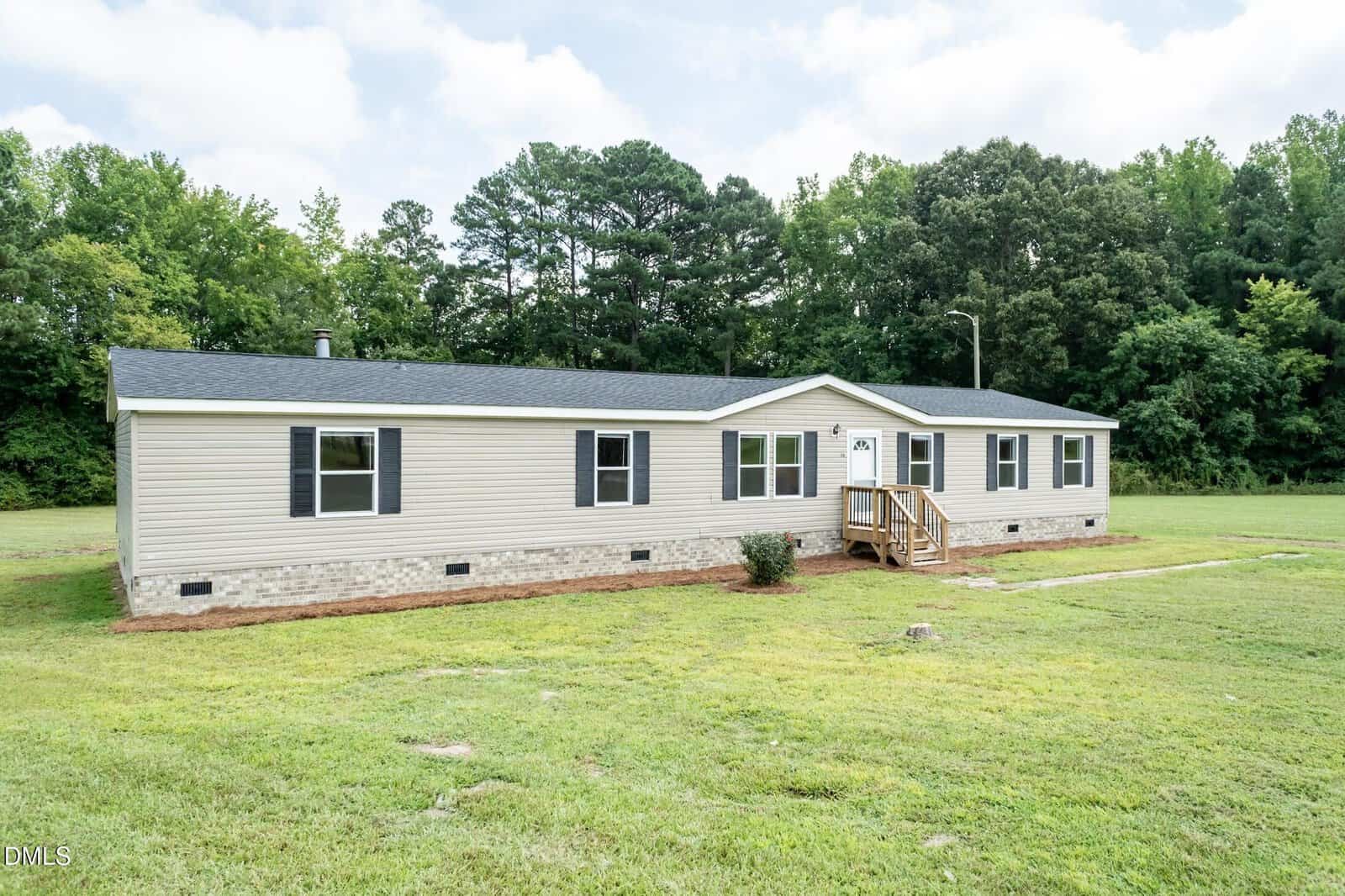 A beige, single-story manufactured home with black shutters and a small front porch sits on a grassy lawn, with trees and a cloudy sky in the background.