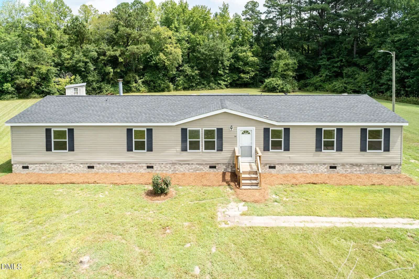 A beige double-wide manufactured home with black shutters, a small front porch with steps, and a large grassy yard sits at 50 Beaver Run in Spring Hope, set against a backdrop of green trees and blue sky.