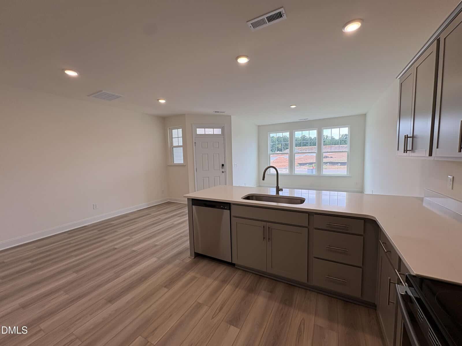 Bright modern kitchen with light wood floors, beige cabinets, white countertops, built-in sink and dishwasher, open to a living area with large windows and a front door. Recessed ceiling lights.