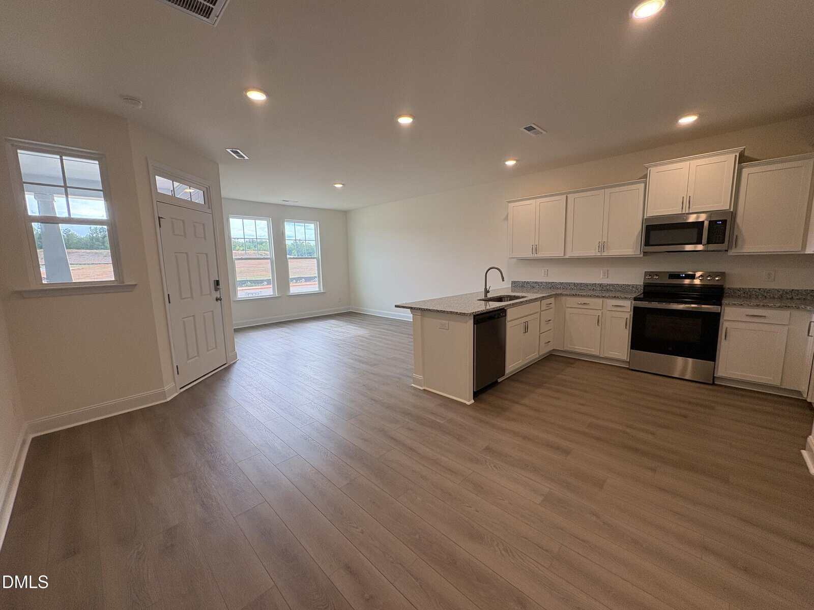 Open modern kitchen with white cabinets and stainless steel appliances next to a spacious living area with large windows, wood floors, and a door leading outside.
