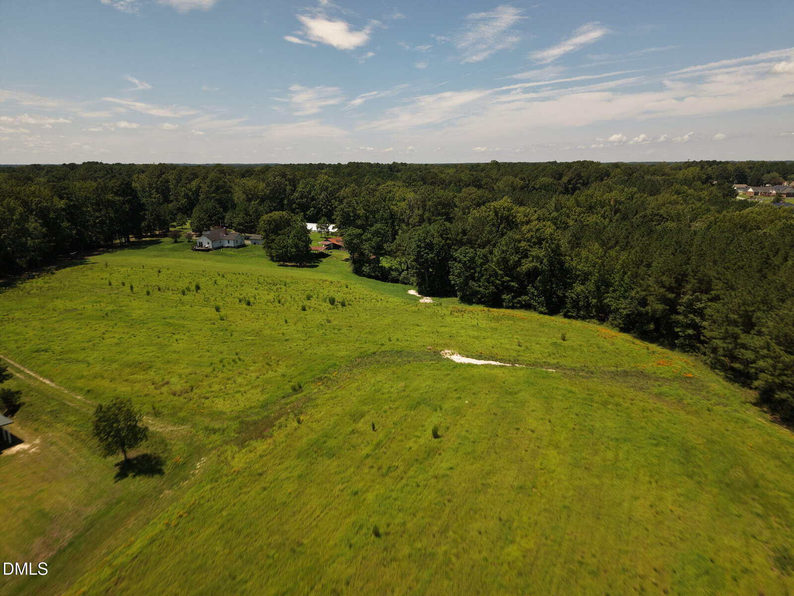 A large green field on tract B along Brassfield Road in Creedmoor, bordered by trees, with a few houses scattered in the distance under a partly cloudy blue sky.