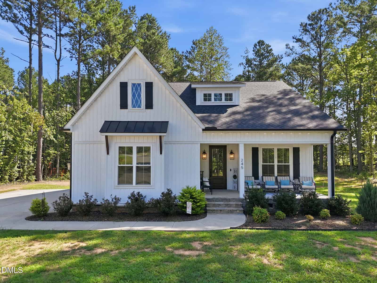 A charming modern farmhouse with white vertical siding, black shutters, and a dark roof. The home features a covered front porch with seating, a stone entryway, and a lush, wooded background under a bright blue sky.