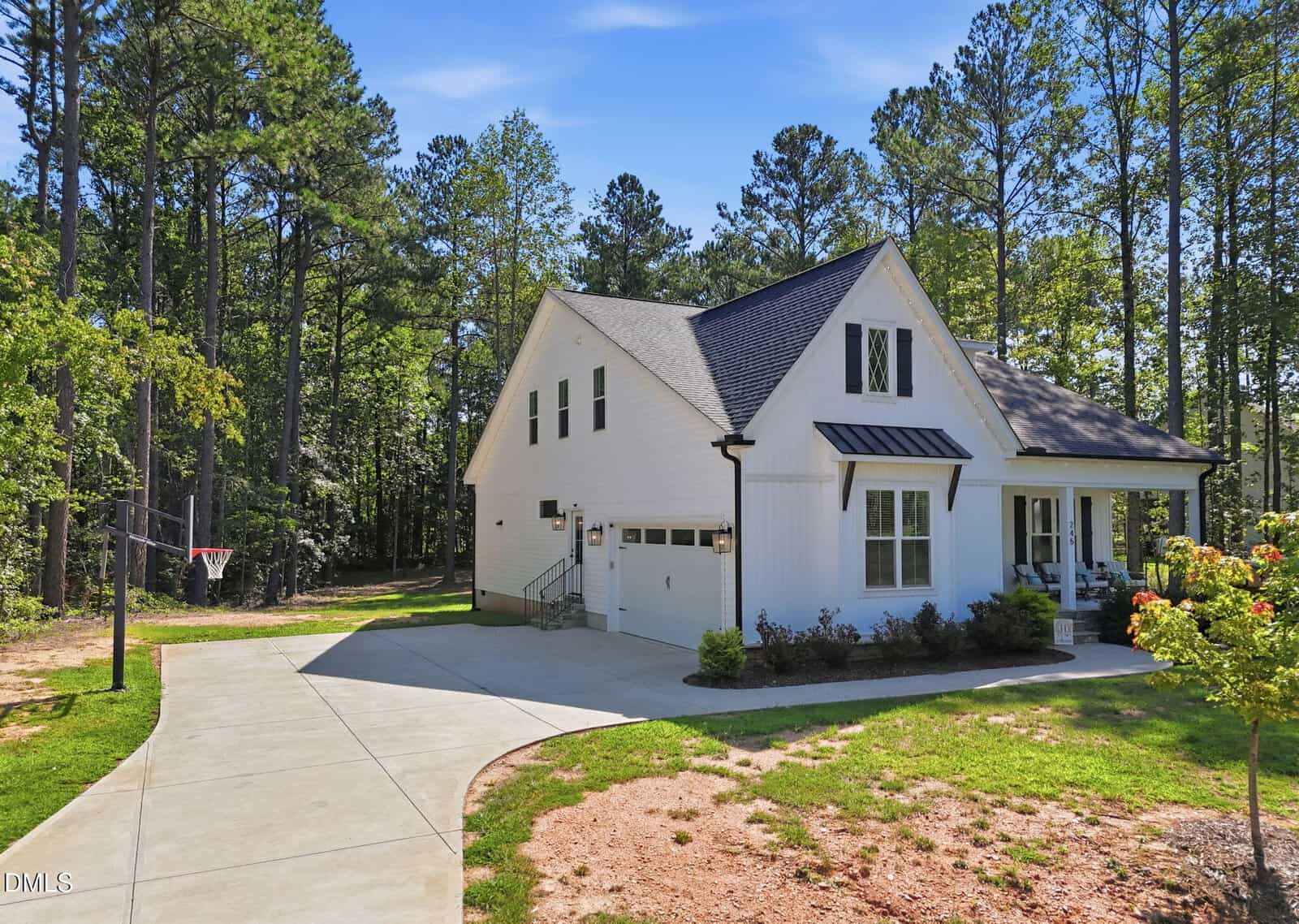 A two-story white farmhouse with black trim and a dark roof, this charming home at 245 Purslane Drive in Franklinton features a single-car garage and a welcoming front porch. A concrete driveway leads to the garage, while a basketball hoop stands on the lawn surrounded by tall pine trees under a blue sky.