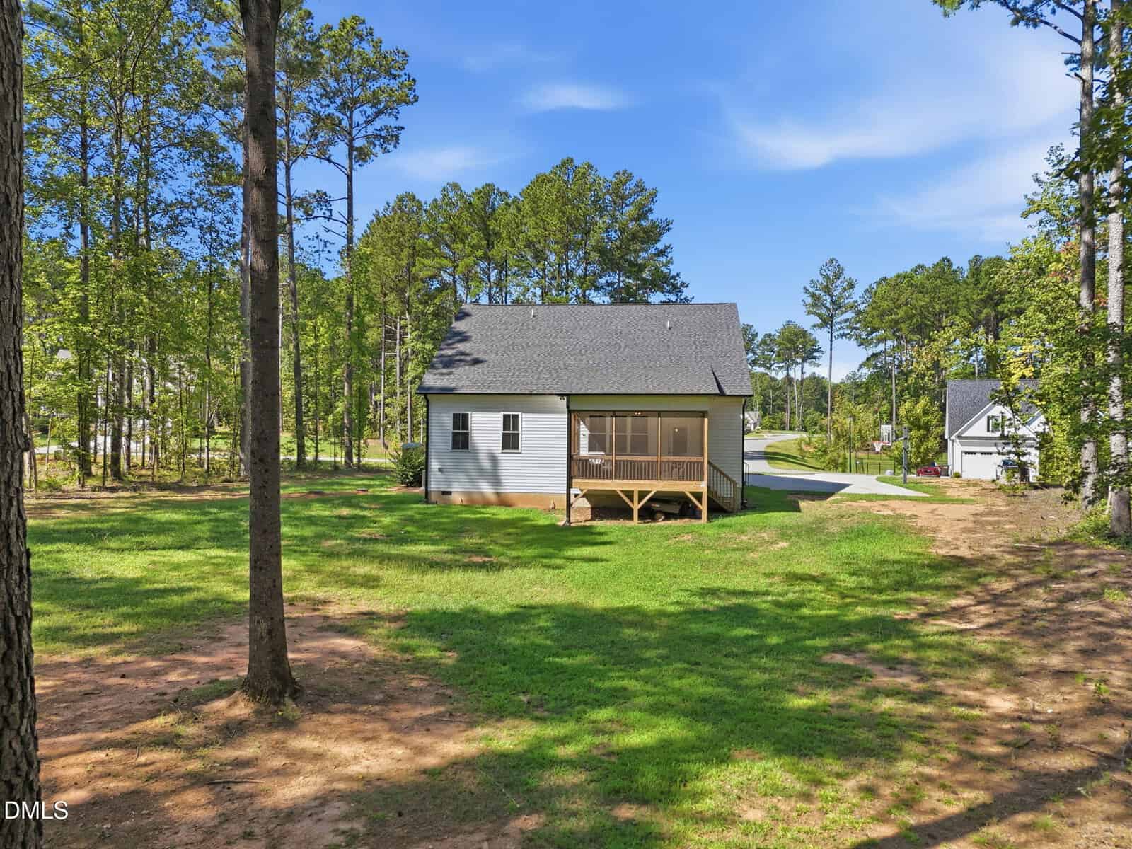 A small, gray house with a porch sits on a grassy lot surrounded by tall trees, with a clear blue sky and another house visible in the background.