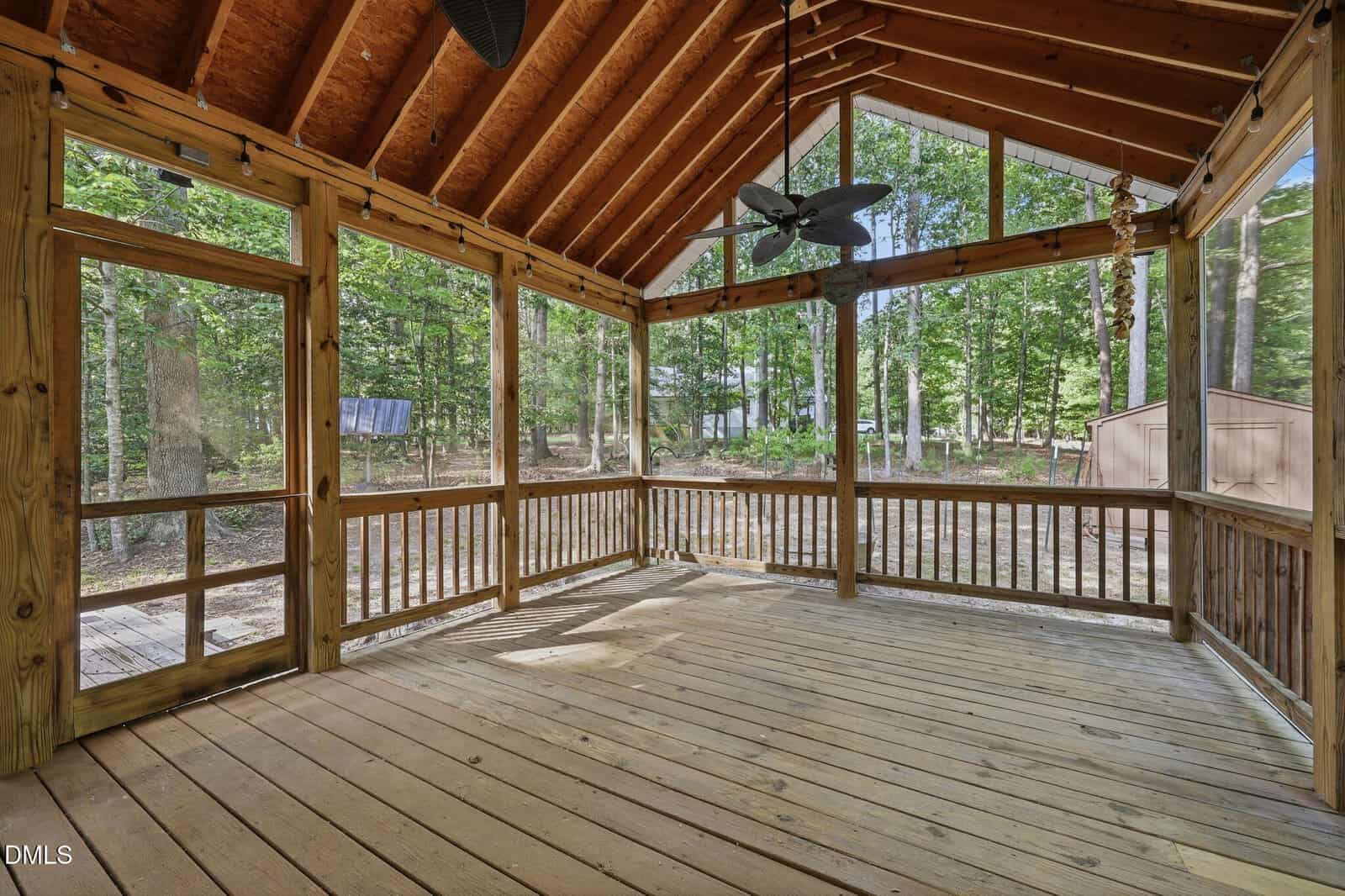 Screened-in wooden porch with a vaulted ceiling, ceiling fan, and views of a wooded backyard with trees and a shed. Sunlight filters through the large screened windows onto the deck floor.