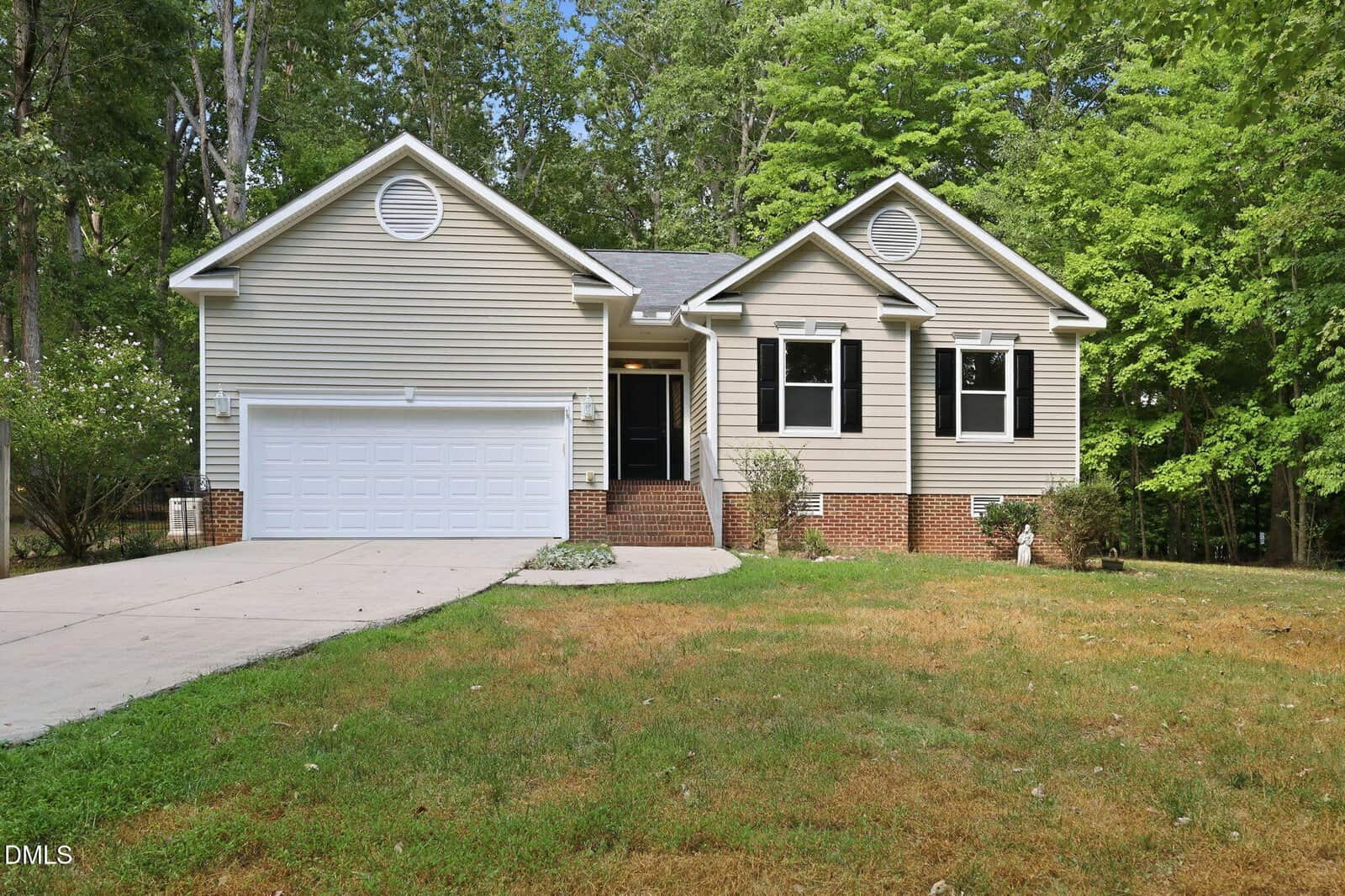 Single-story house with beige siding, white trim, two front gables, a two-car garage, brick foundation, black front door, small steps, and a lawn with some patches and shrubs, surrounded by tall trees—all nestled on 1268 Sagamore Drive in the quiet community of Louisburg.