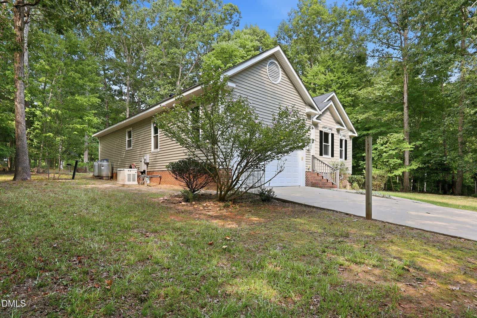 Single-story beige house with white trim, steps leading to the front door, surrounded by trees and grass, with a concrete driveway and visible outdoor HVAC units on the left side.