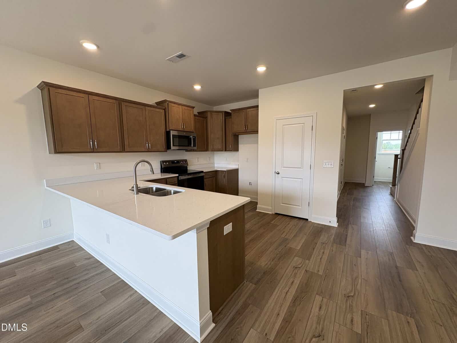 Modern kitchen with brown cabinets, white countertops, stainless steel appliances, sink on an island, and wood-style flooring. A hallway and staircase are visible in the background. Bright lighting with recessed ceiling lights.