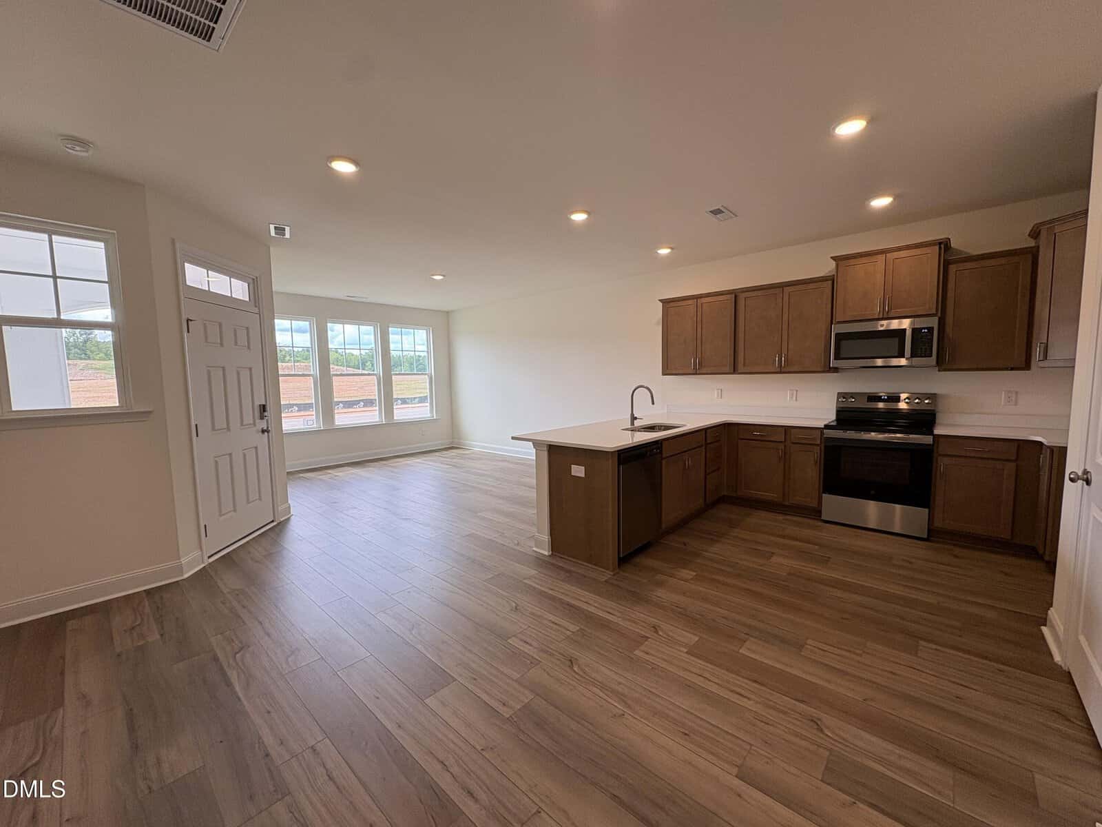 Open-concept living space with wood-look flooring, a modern kitchen with brown cabinets and stainless steel appliances, large windows, and a white front door. Recessed lighting brightens the room.