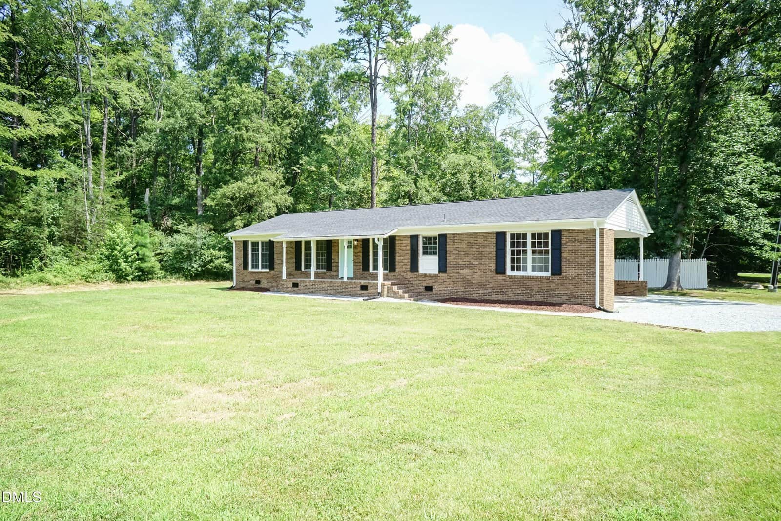 Single-story brick house with white trim and black shutters, set on a spacious grassy yard with trees in the background under a sunny sky.