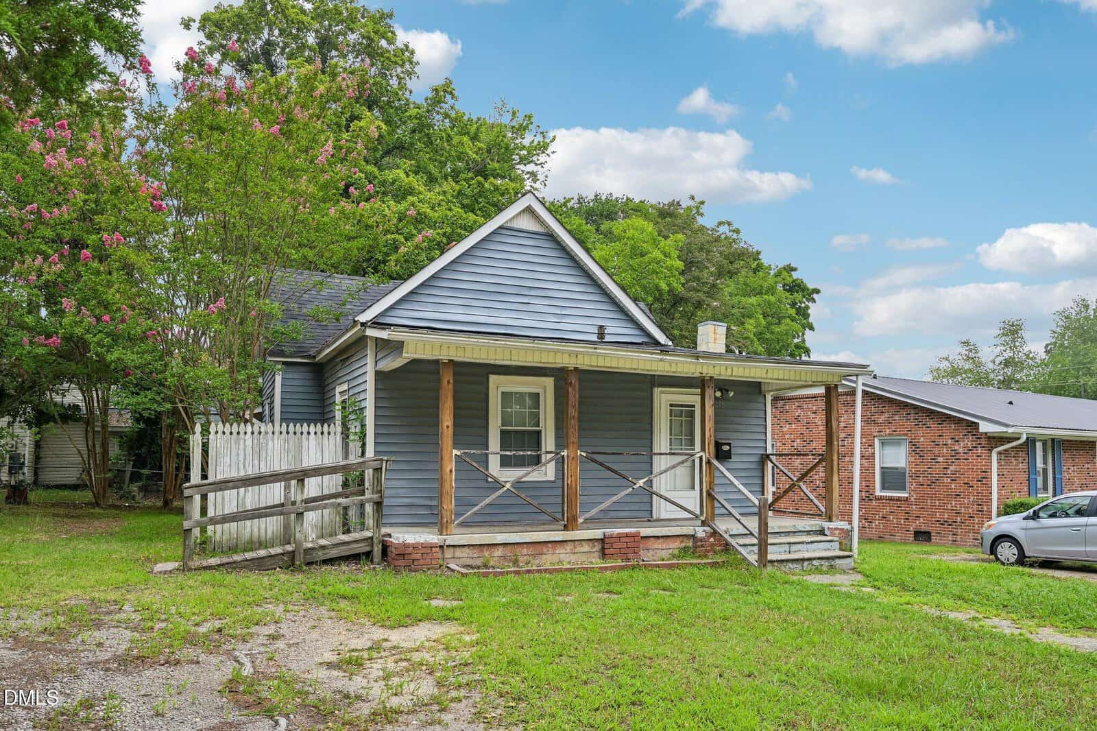 A small blue house with a covered front porch, wooden railing, ramp, and fenced yard is surrounded by green trees and neighboring brick homes under a blue sky with clouds.