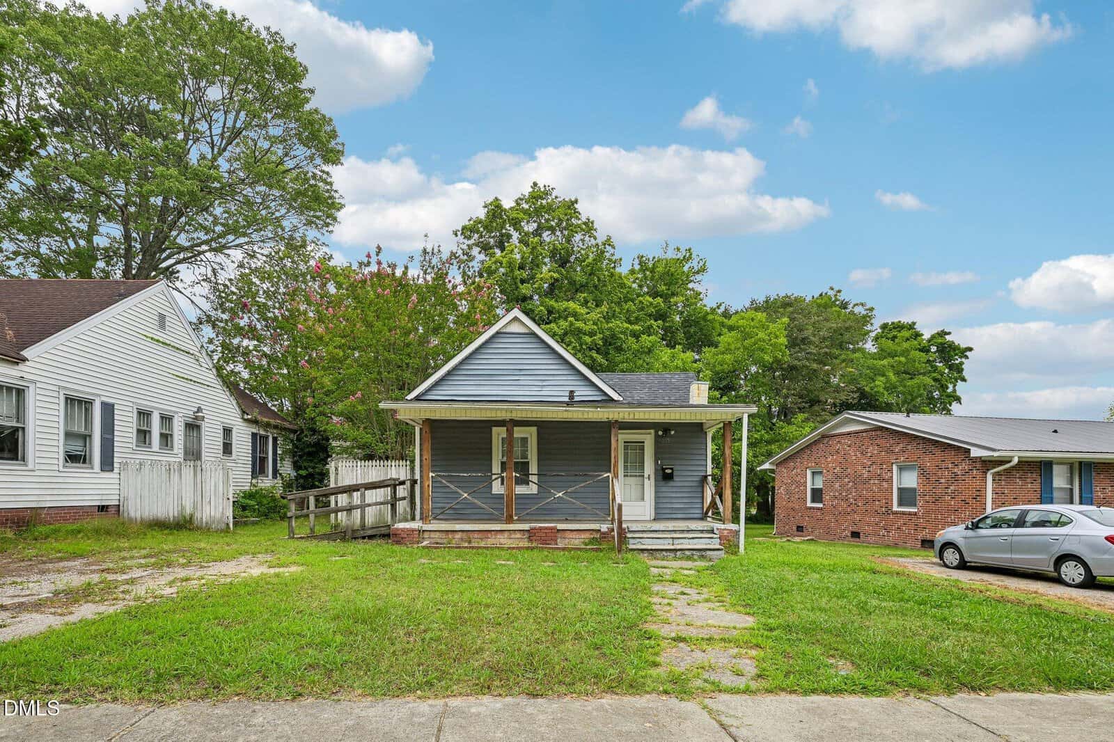 Small blue house with a front porch and ramp, flanked by neighboring homes, sits on a grassy lot under a partly cloudy sky. A silver car is parked next door.