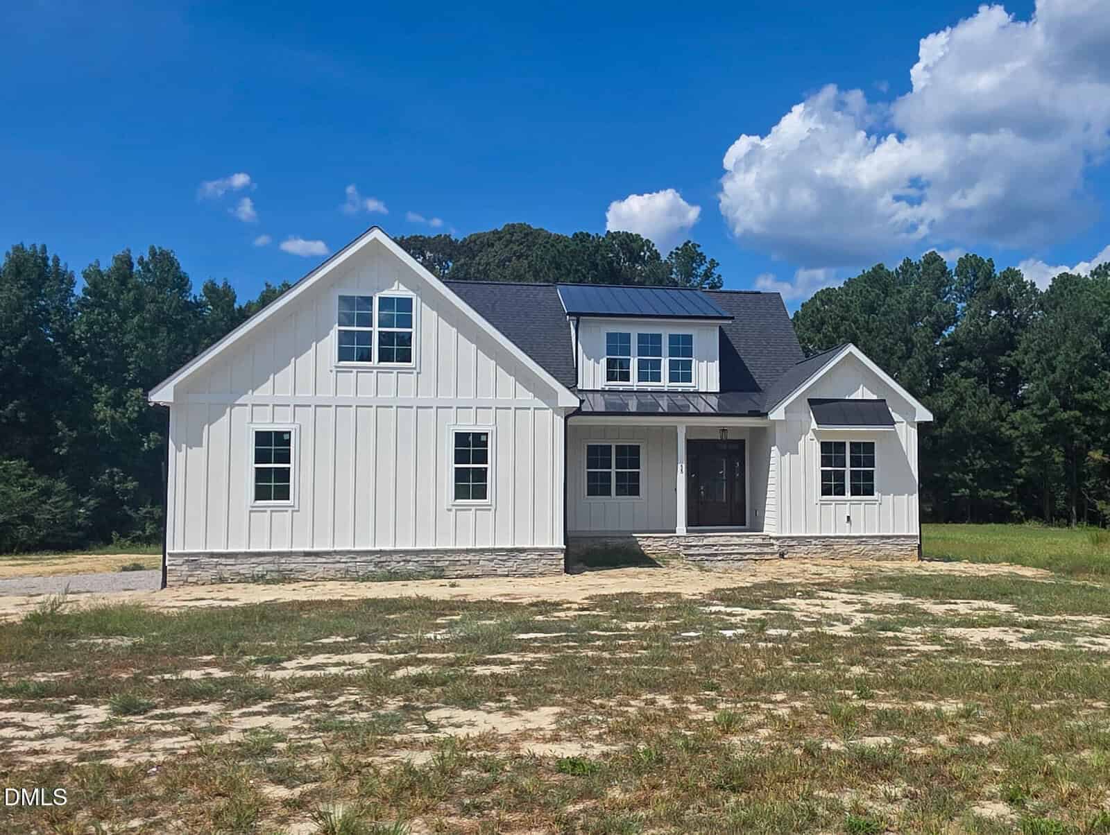 Modern white farmhouse with black roof and stone accents, set on a grassy lot with some bare patches, surrounded by trees under a blue sky with scattered clouds—this stunning property at 55 White Deer Lane in Youngsville offers timeless charm and contemporary appeal.
