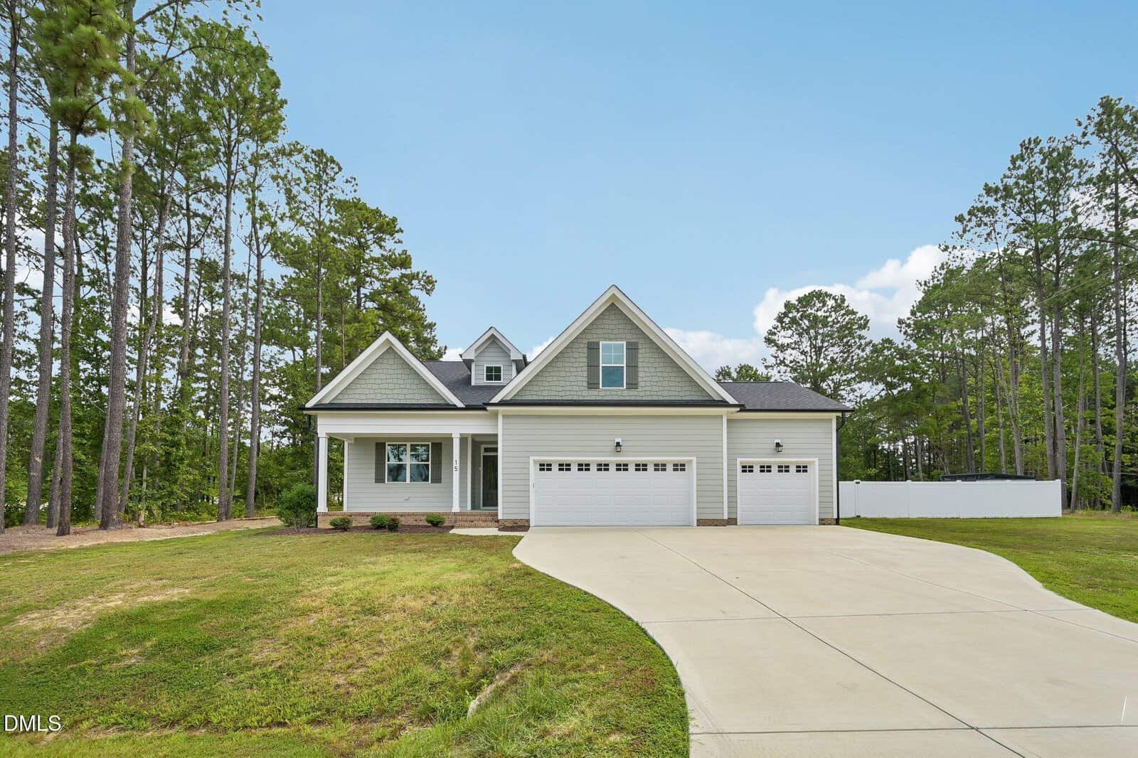 A modern, light gray house with a three-car garage and a large driveway sits among tall pine trees, with a well-kept lawn and a white fence on the right.