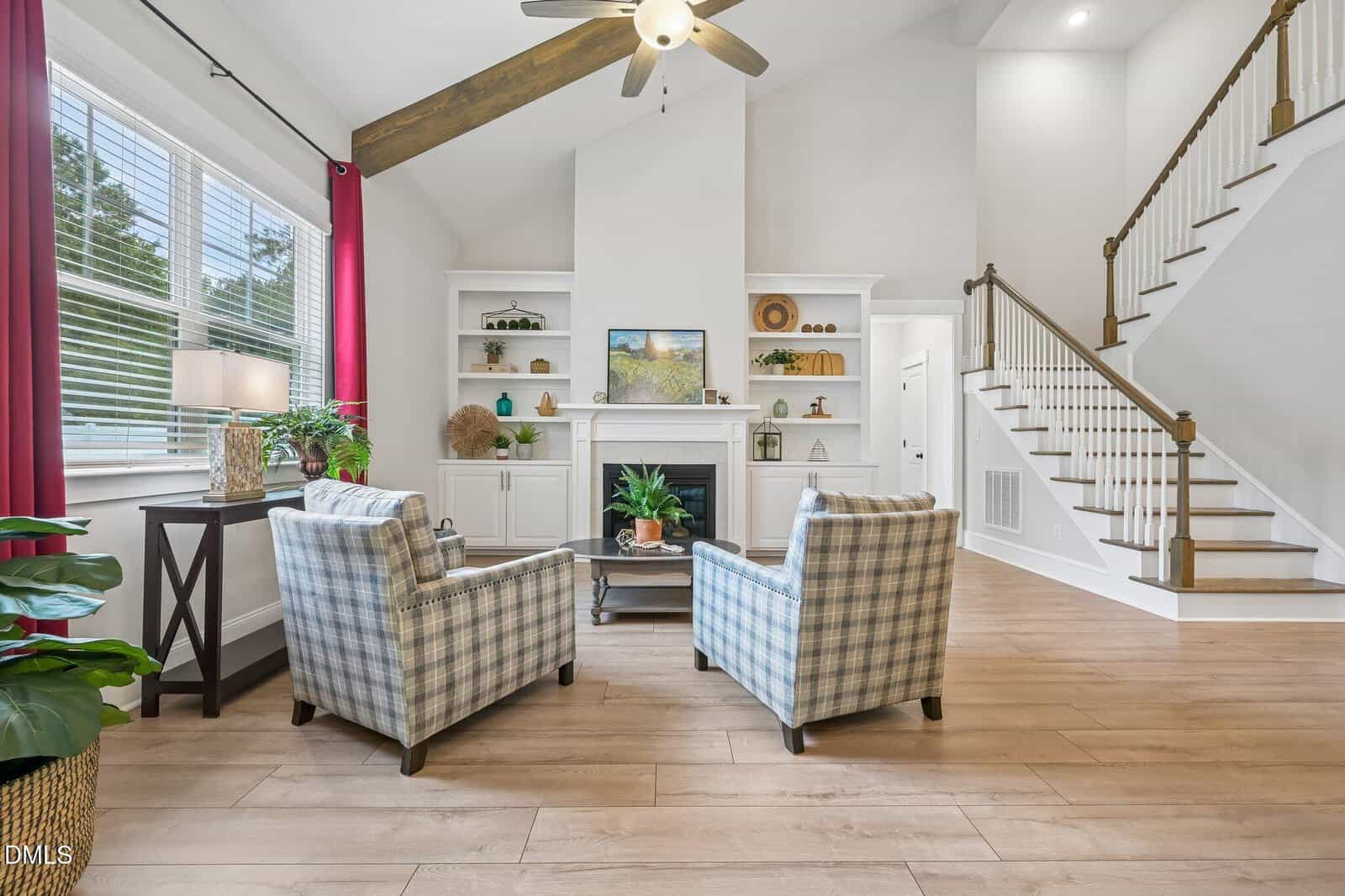 Bright living room with two plaid armchairs facing a fireplace, built-in shelves, potted plants, wood floors, large windows with red curtains, and a staircase to the right.