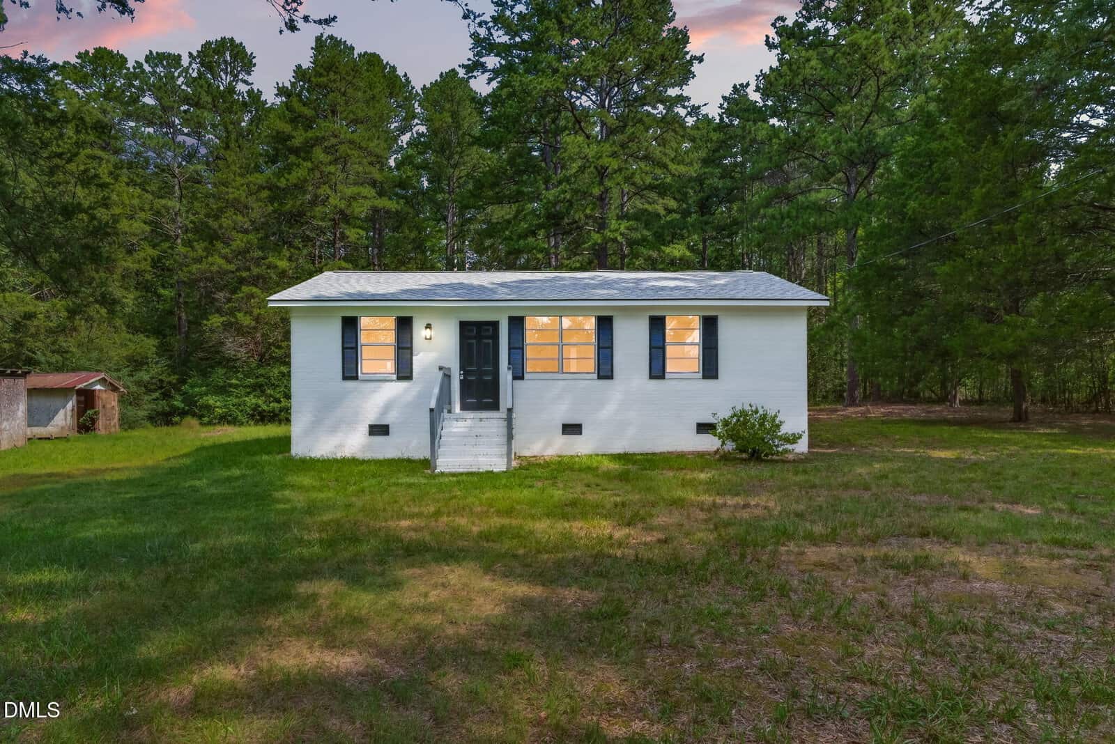 A small white house with blue shutters, lit windows, and a front stoop stands in a grassy clearing surrounded by tall trees at dusk—a charming scene reminiscent of Bullock’s homestead at 3629 Plantation Road.